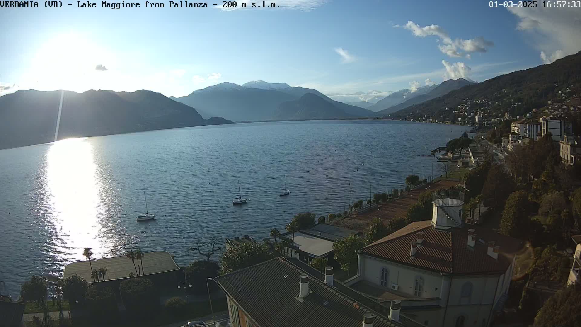 A sunny day overlooking a calm lake with mountains in the background, showing a shoreline town with buildings and tennis courts.