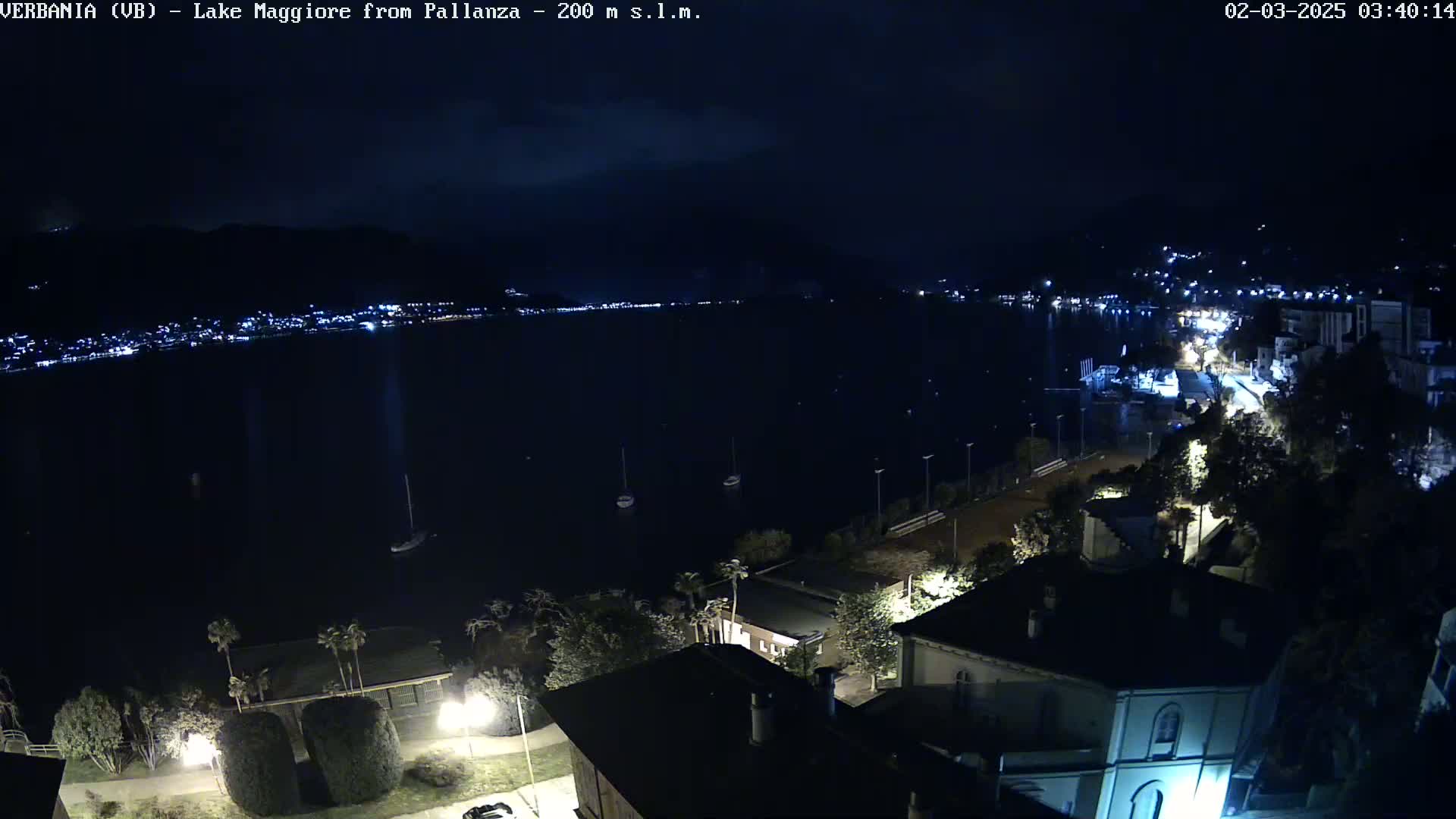 A nighttime view of a calm lake with lights from a town visible on the far shore, and buildings and trees illuminated in the foreground.