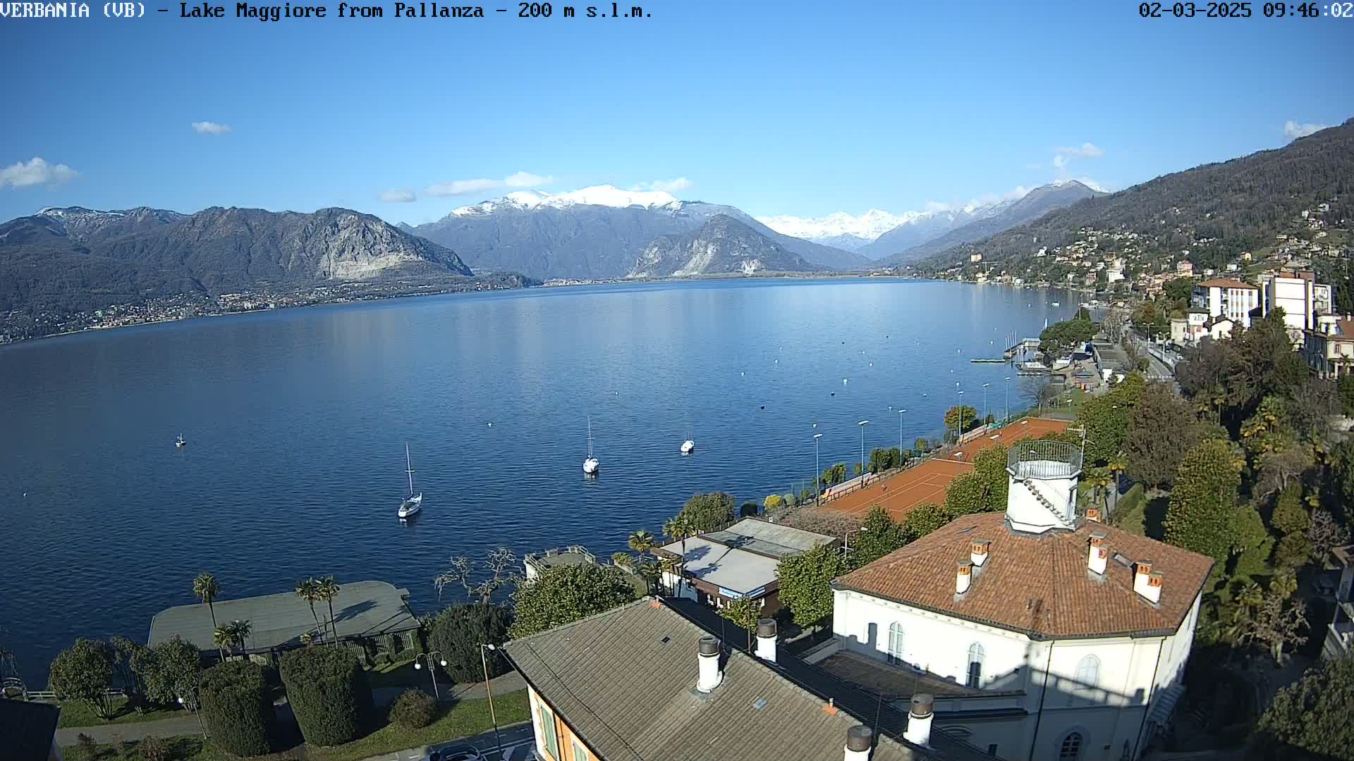 A picturesque view of Lake Maggiore from Pallanza, on a sunny day with snow-capped mountains in the background, shows sailboats on the water and buildings along the shoreline.