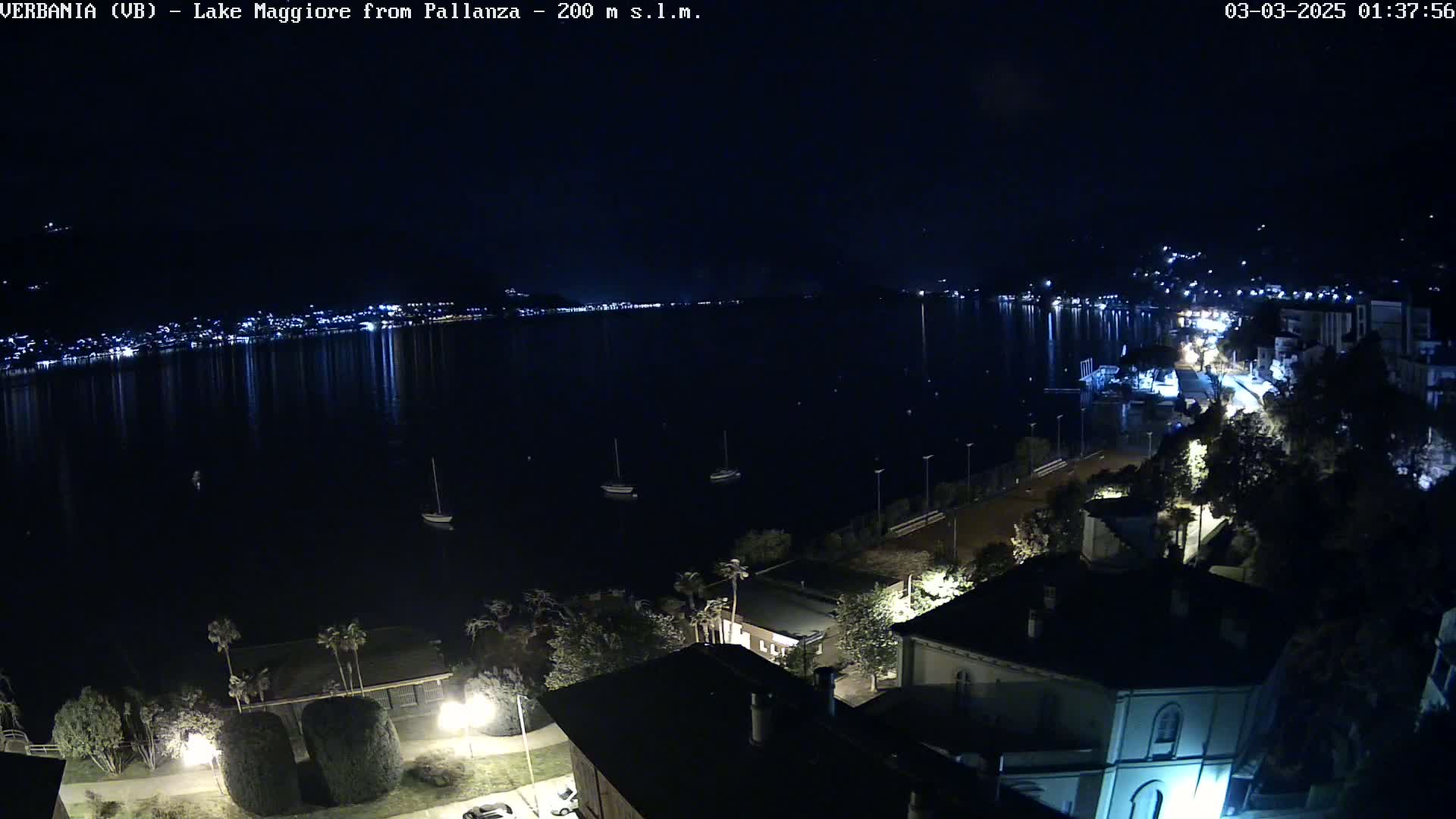 A nighttime view of a calm lake reflecting the lights of a town across the water, with a few boats visible and buildings and trees in the foreground.