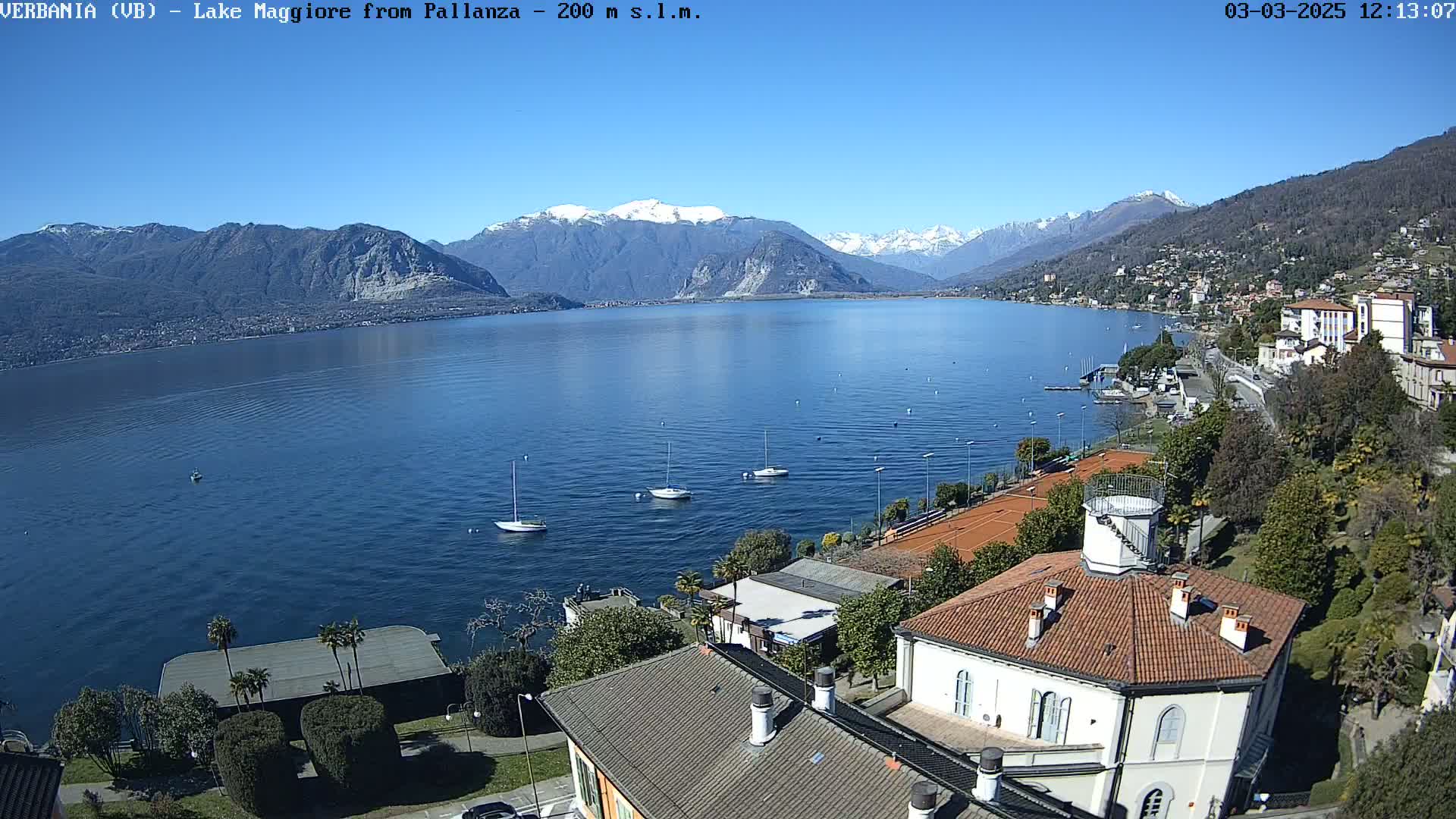 A clear, sunny day reveals a lake with several sailboats, a shoreline with buildings and tennis courts, and snow-capped mountains in the background.