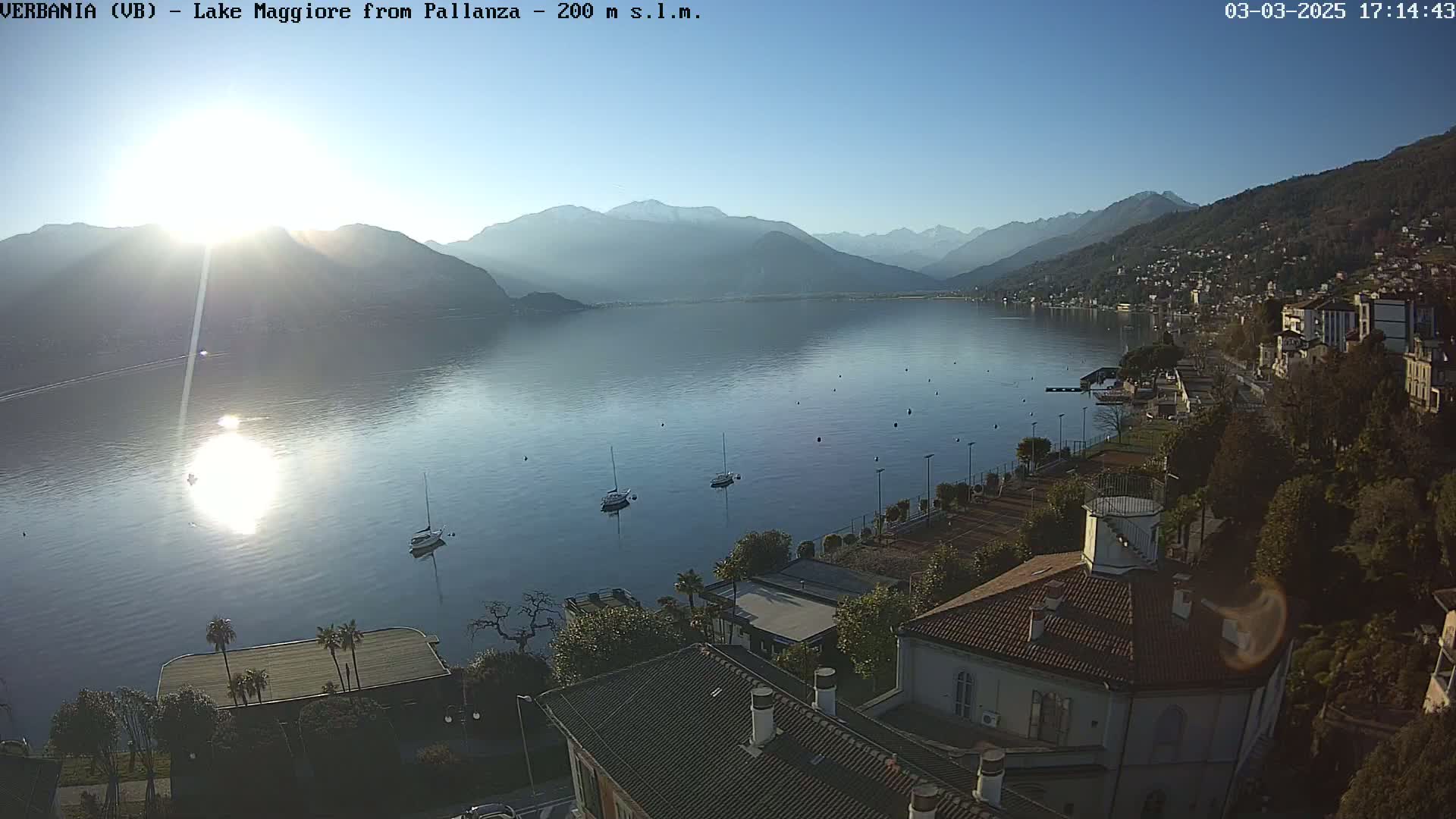 A sunny, clear day overlooks a calm lake with mountains in the background, several sailboats on the water, and buildings and trees along the shoreline.