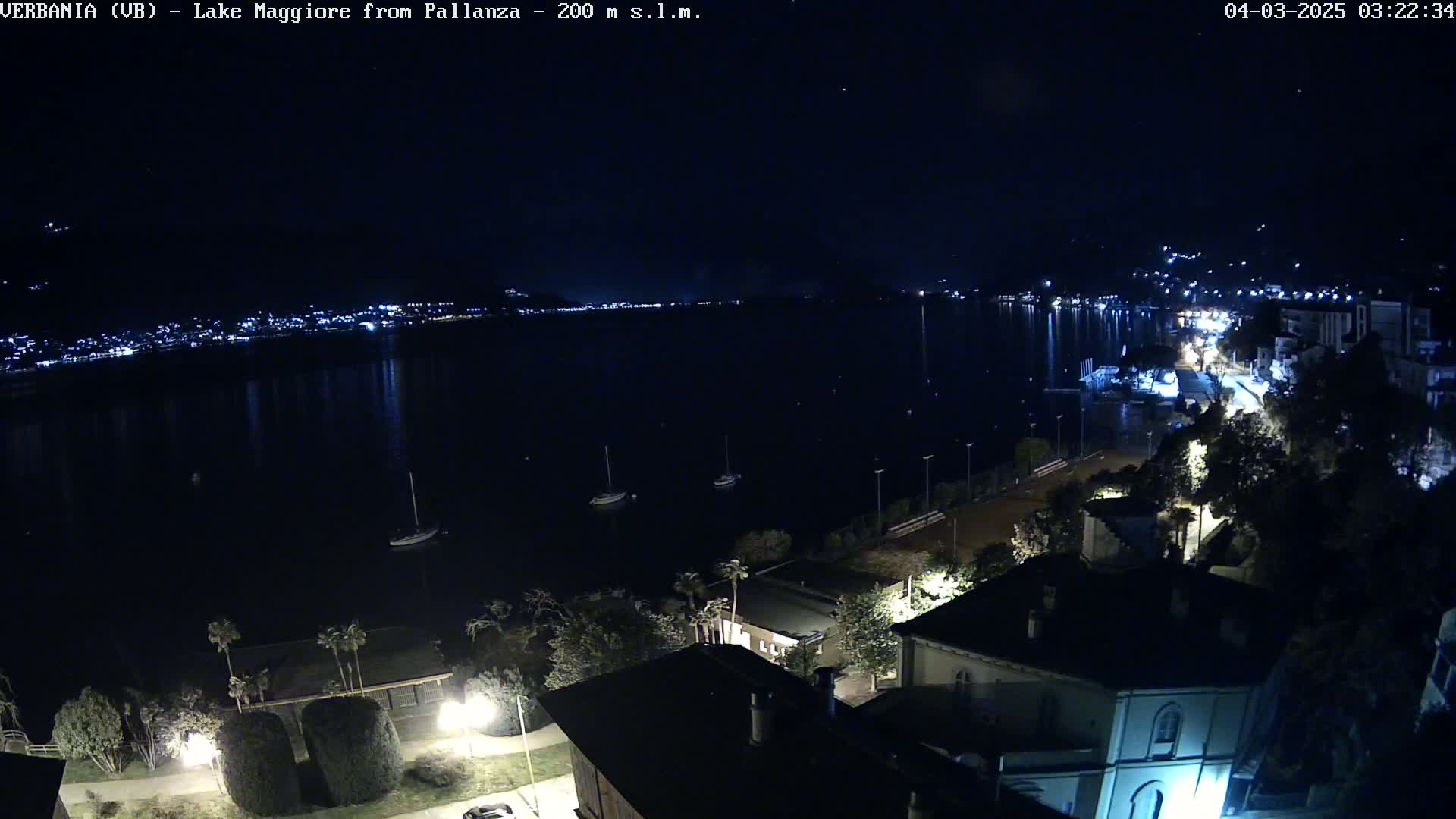 A nighttime view of a calm lake with city lights across the water, a few boats, and buildings and trees on the foreground shore under a clear, dark sky.