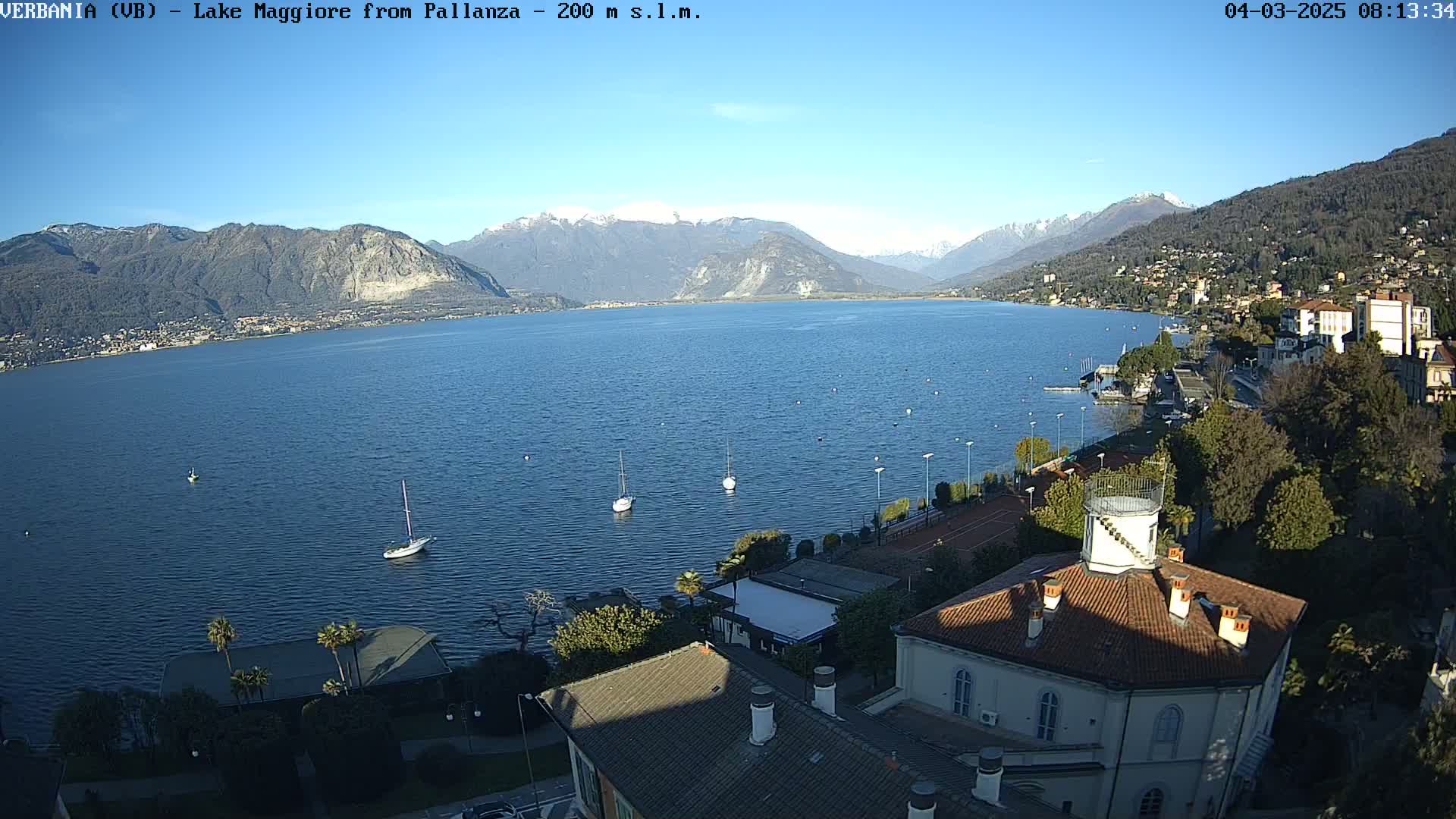 A clear, sunny day overlooking a large lake with sailboats, mountains in the background, and buildings along the shoreline.