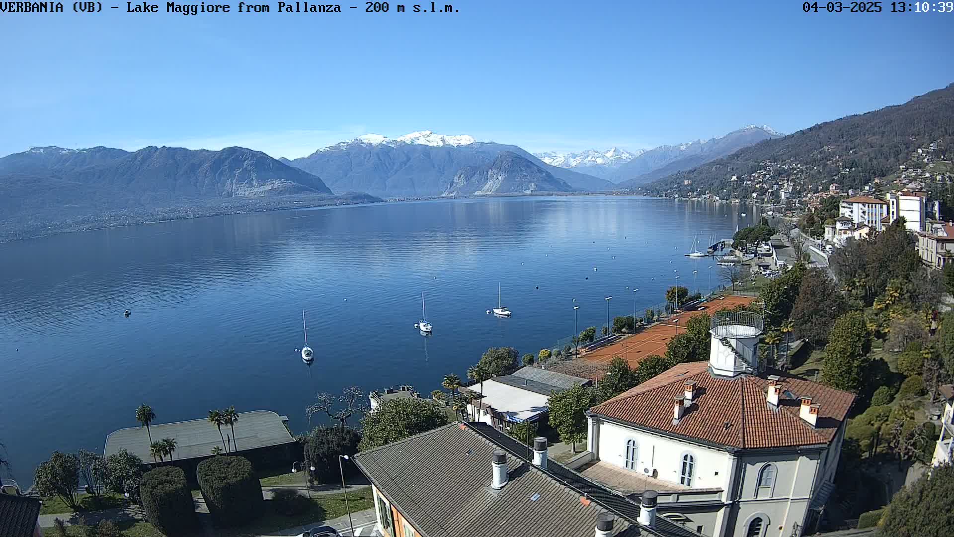 A clear, sunny day overlooks a calm lake with several sailboats and mountains in the background, showcasing a waterfront scene with buildings and tennis courts.