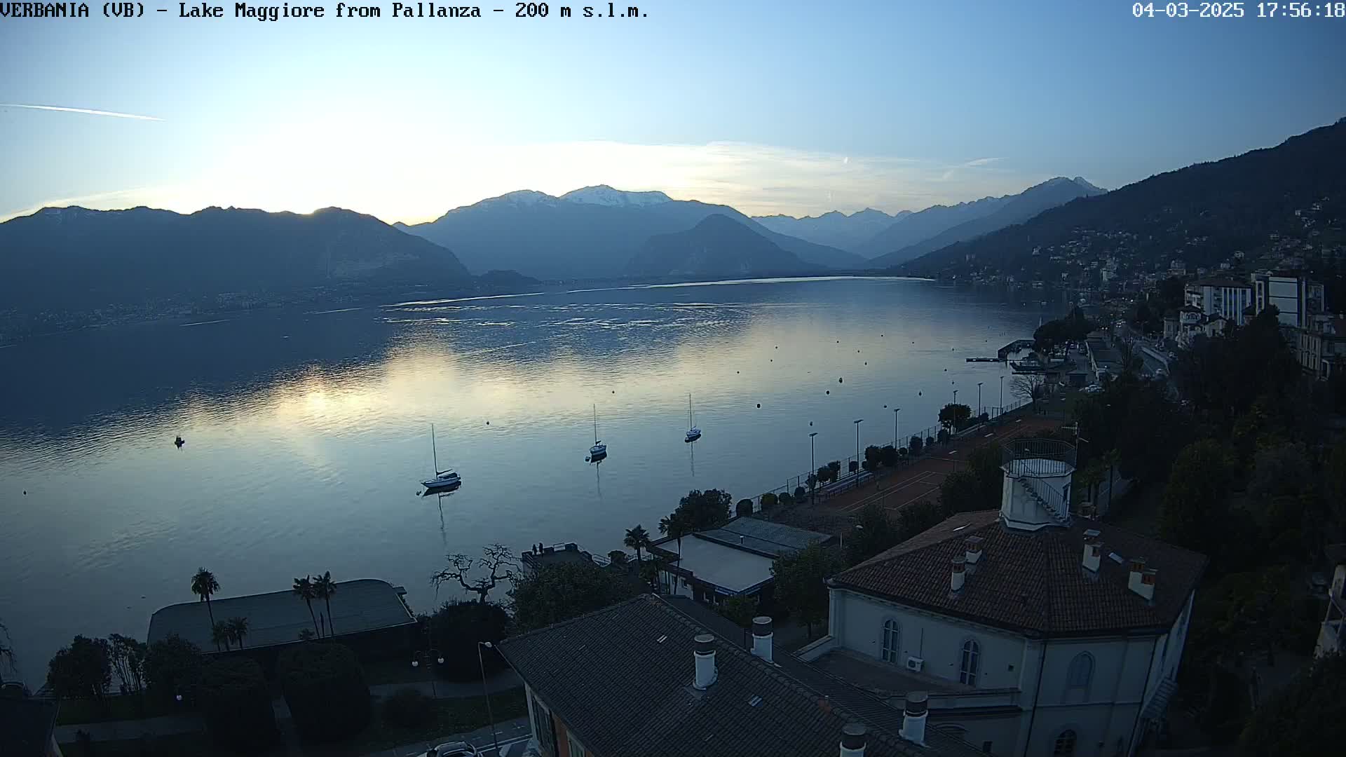A tranquil lake at sunset, reflecting the light against a backdrop of mountains, is viewed from above a lakeside town with several boats on the water.