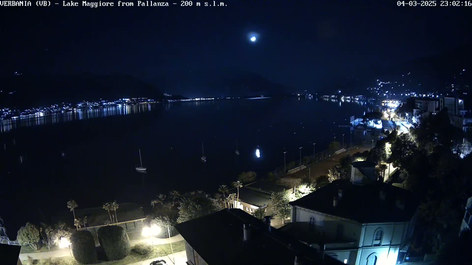 A nighttime, high-angle view of a moonlit lake with a town's lights reflecting on the water, a few boats, and buildings along the shoreline.