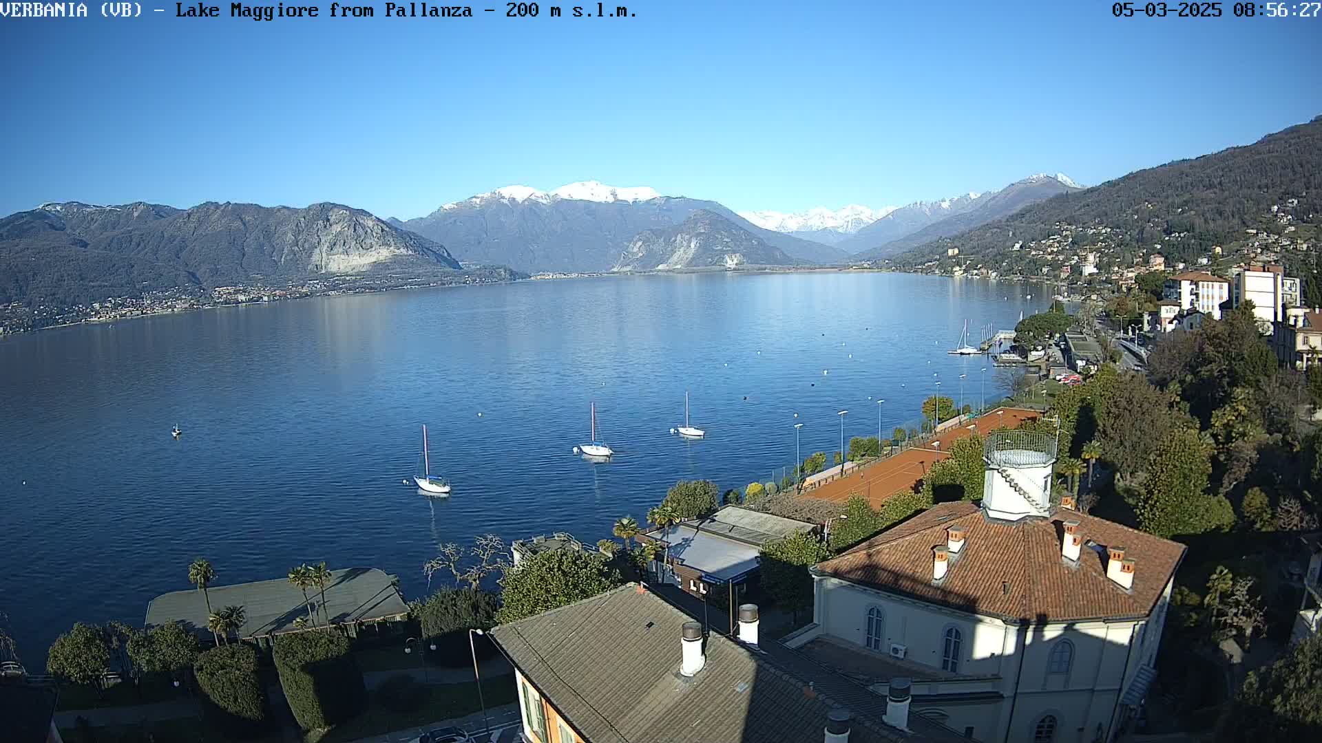 A sunny day overlooking a calm lake with snow-capped mountains in the background, several sailboats on the water, and buildings and tennis courts along the shoreline.