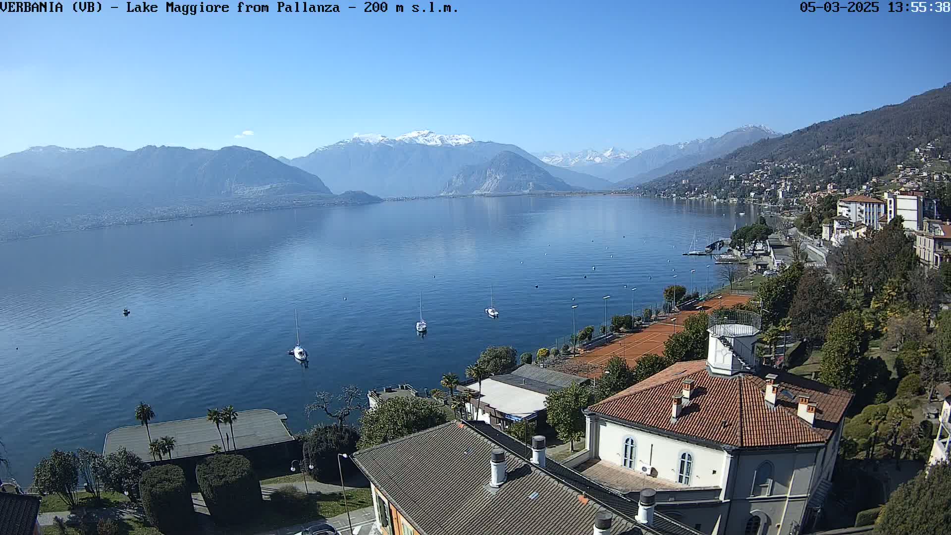 A clear, sunny day overlooks a calm lake with several sailboats, tennis courts, and buildings with red-tiled roofs, backed by snow-capped mountains.