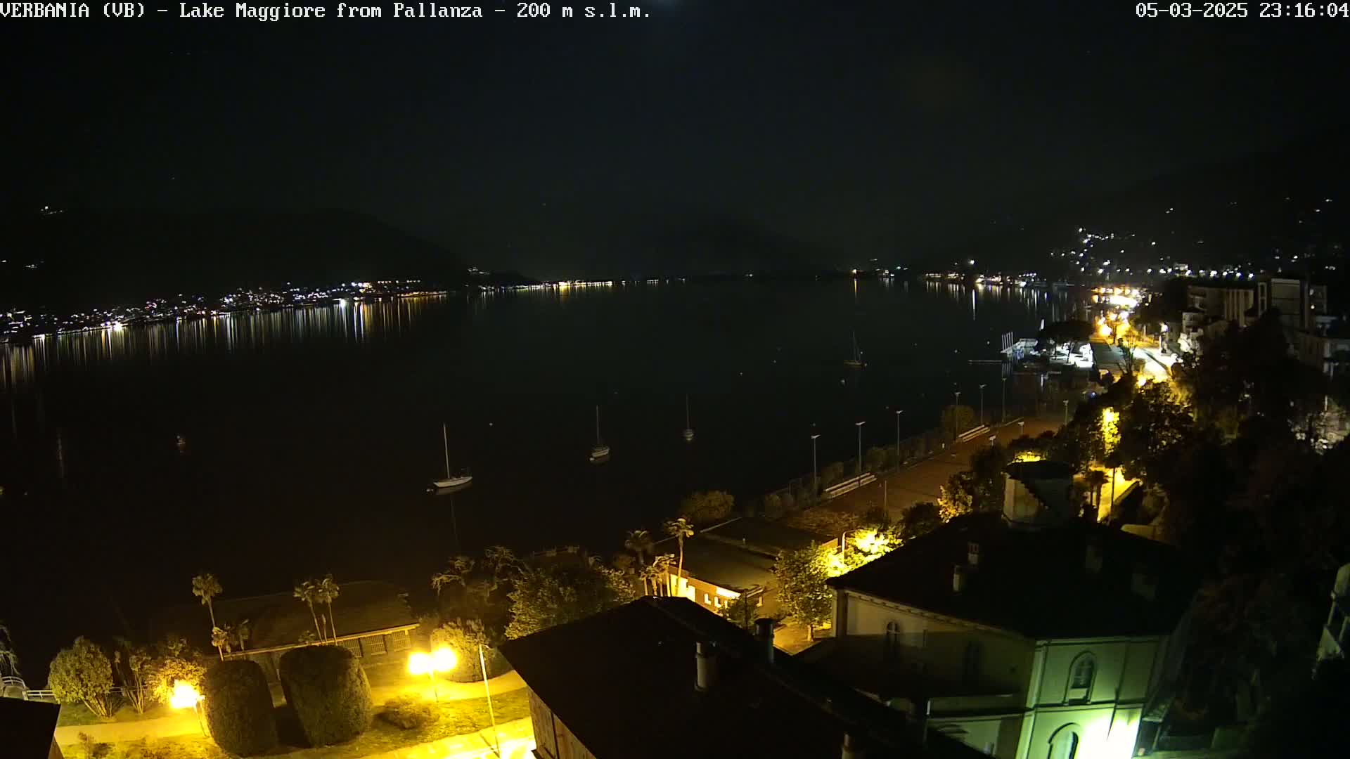 A nighttime view of a calm lake reflecting the lights of a town on its shore, with boats moored in the water and illuminated buildings along the waterfront.