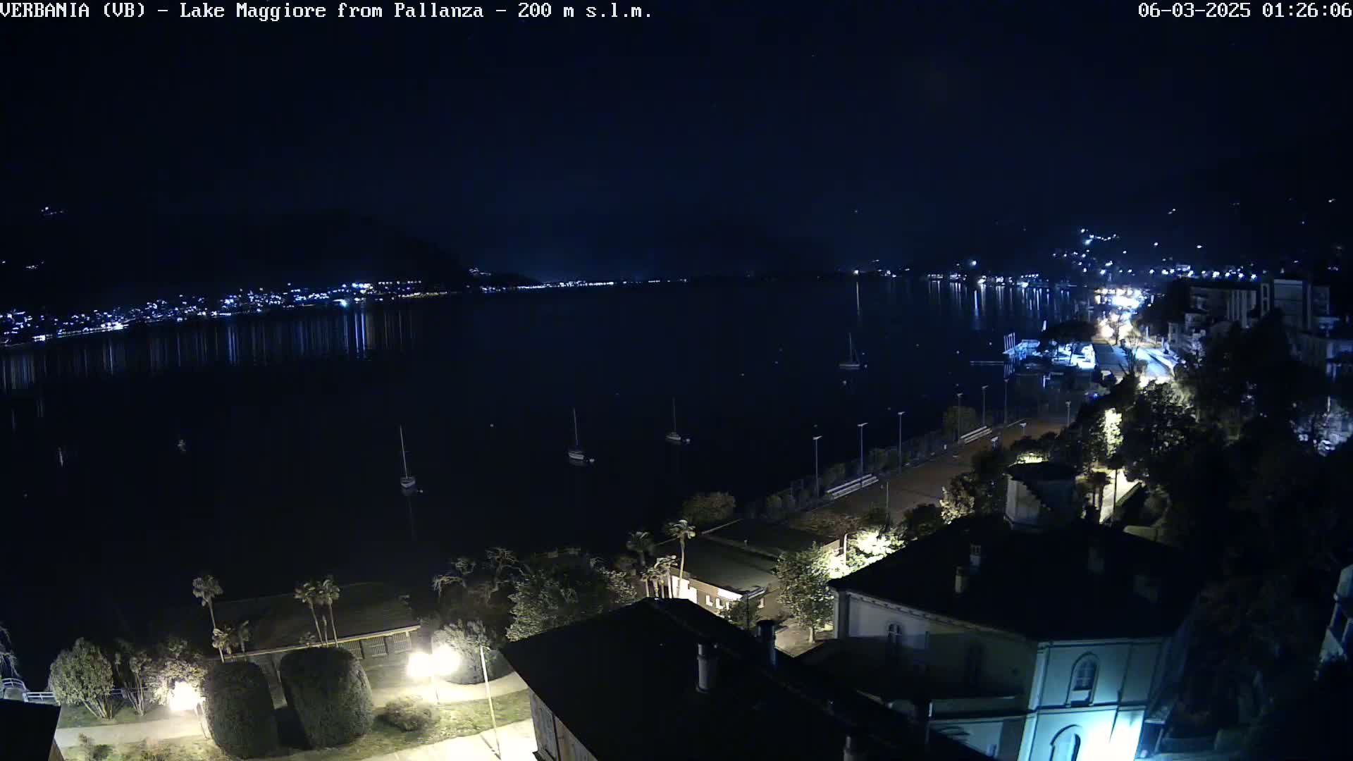 A nighttime view of a calm lake reflecting city lights, with boats moored on the water and buildings illuminated along the shoreline under a clear, dark sky.