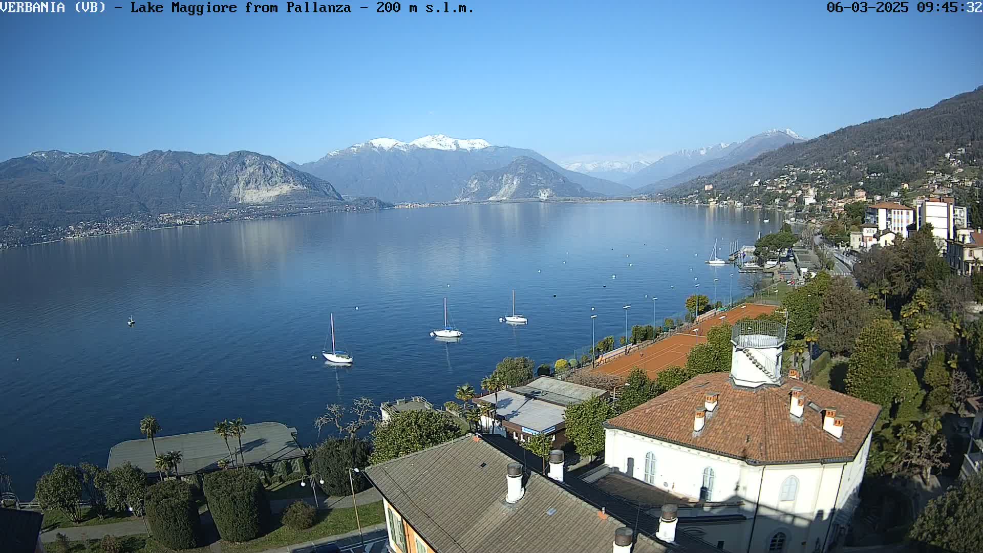A clear, sunny day overlooking a calm lake with several sailboats, tennis courts, and buildings nestled against snow-capped mountains in the background.