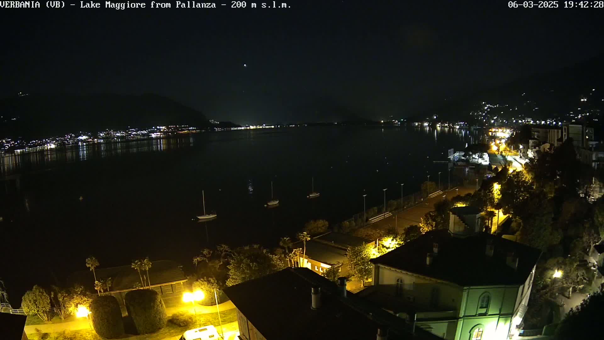 A nighttime, high-angle view of a dark lake with several boats, a town's lights reflecting on the water, and a shoreline with buildings and streetlights.