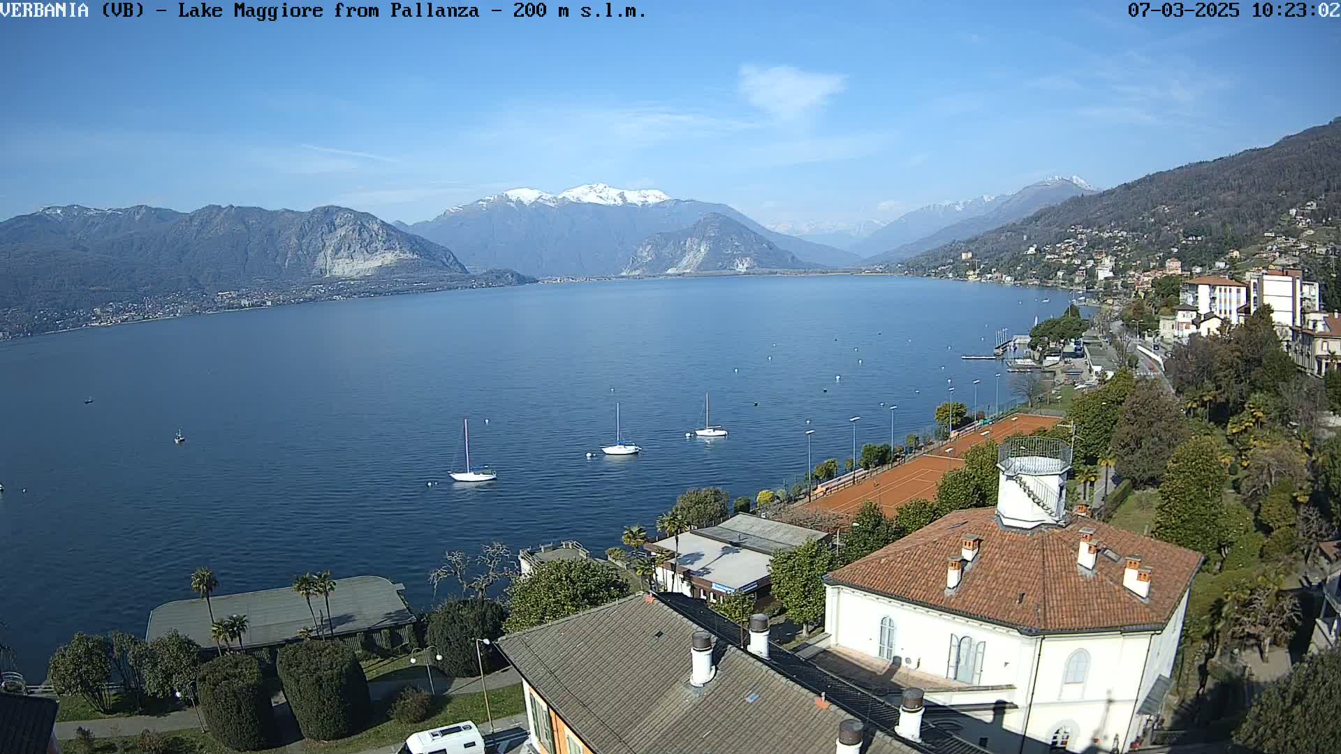 A sunny day overlooking a calm lake with several sailboats, tennis courts, and buildings nestled along the shoreline, mountains in the background showing some snow.