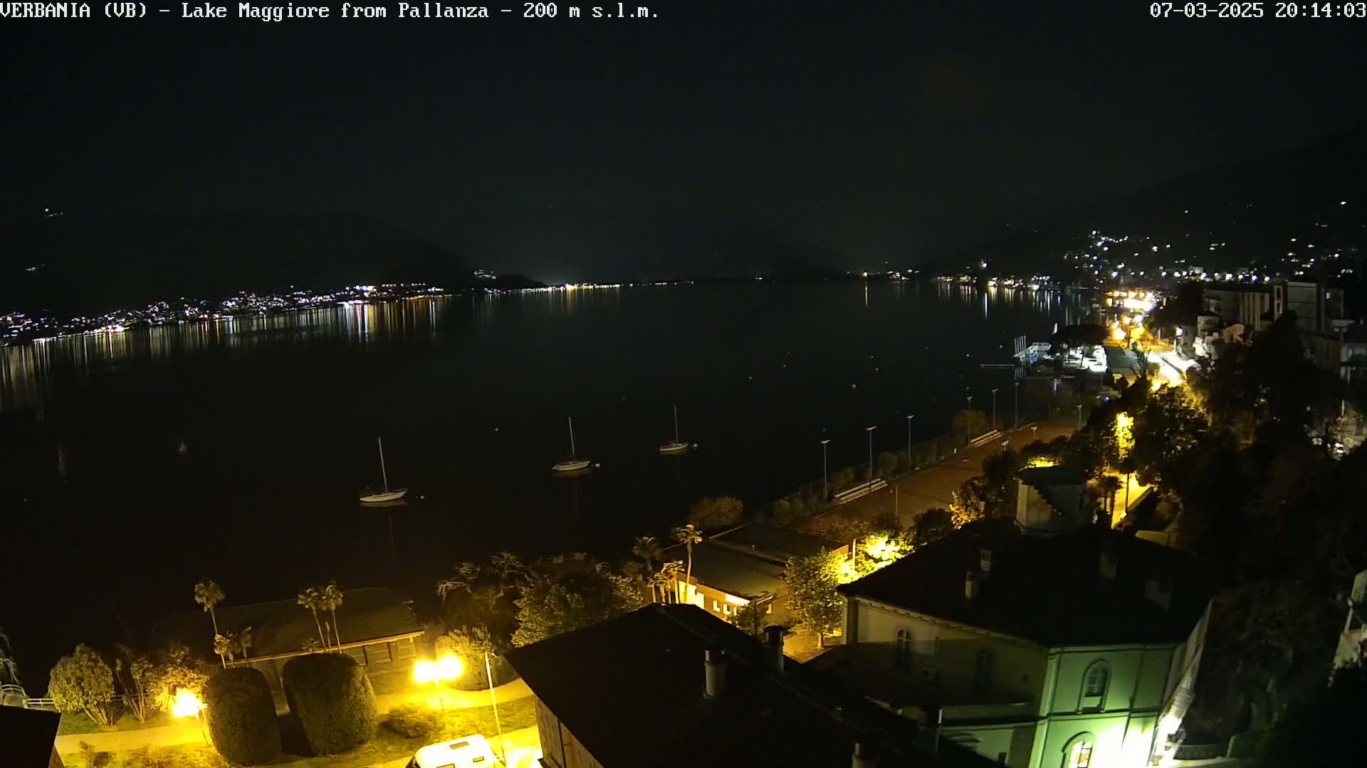 A nighttime view of a calm lake reflecting city lights, with a few sailboats moored, and a shoreline lined with buildings and trees illuminated by streetlights.