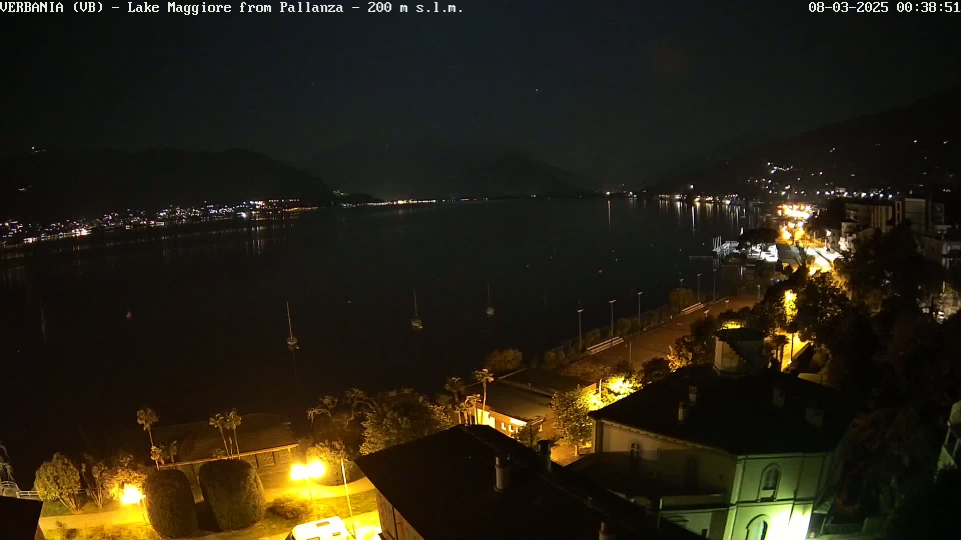 A nighttime view of a calm lake with a few boats, a shoreline with illuminated buildings and trees, and hills in the background under a clear sky.