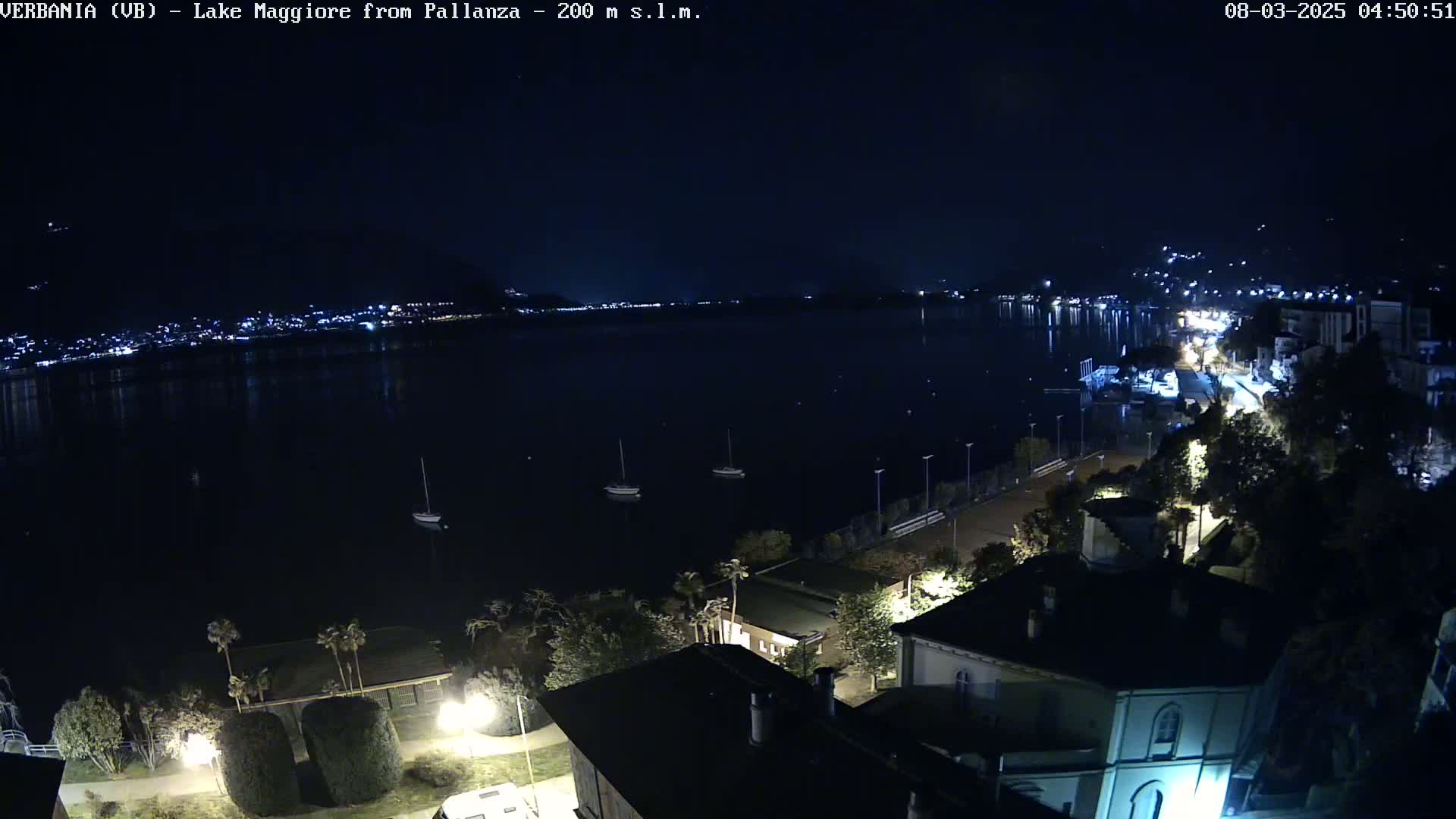 A nighttime, high-angle view shows a dark body of water with a few boats, a town with lights along the shoreline, and some buildings and trees in the foreground.
