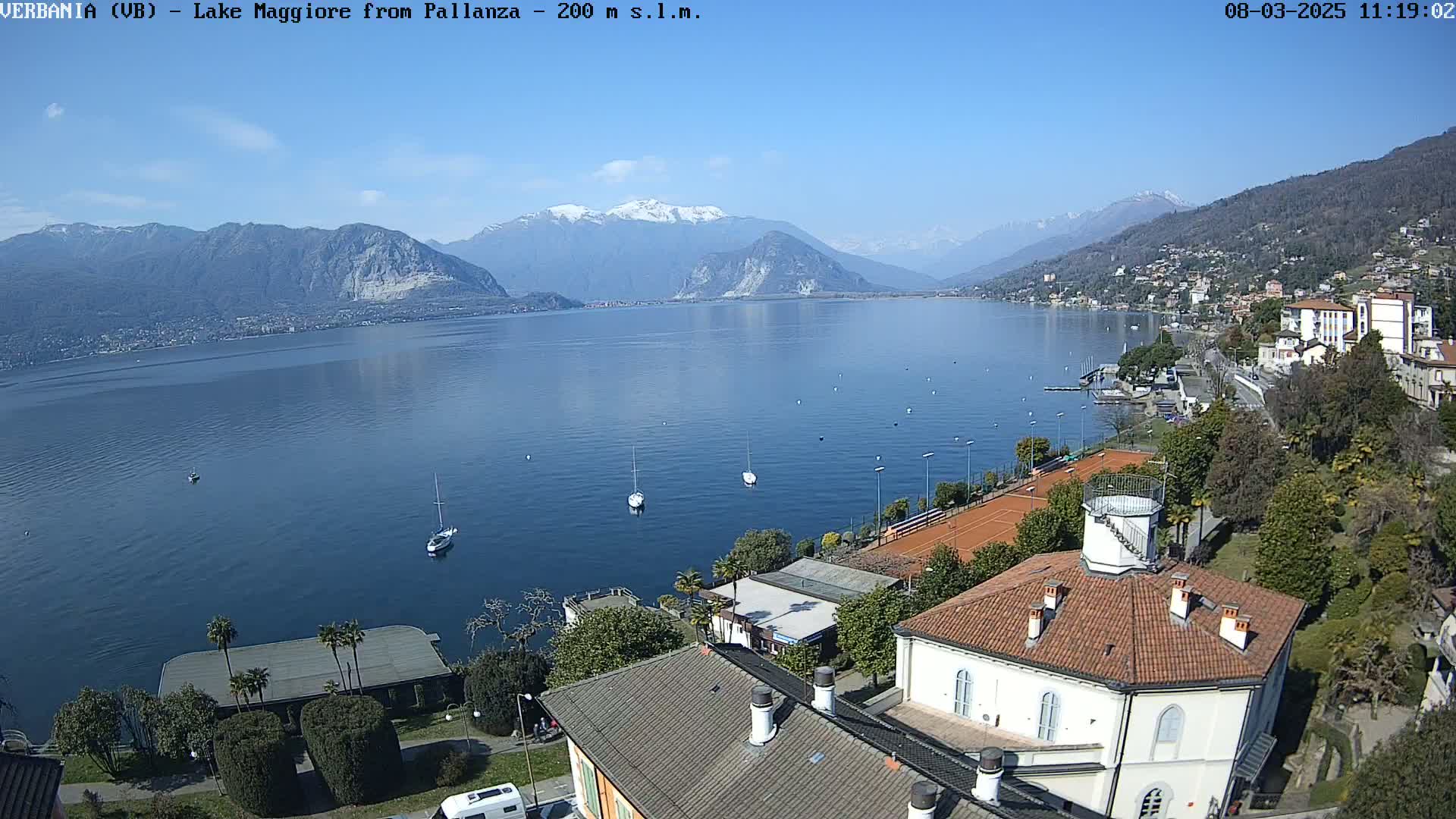 A sunny day overlooking a calm lake with snow-capped mountains in the background, featuring several boats on the water and a coastal town with buildings and tennis courts.