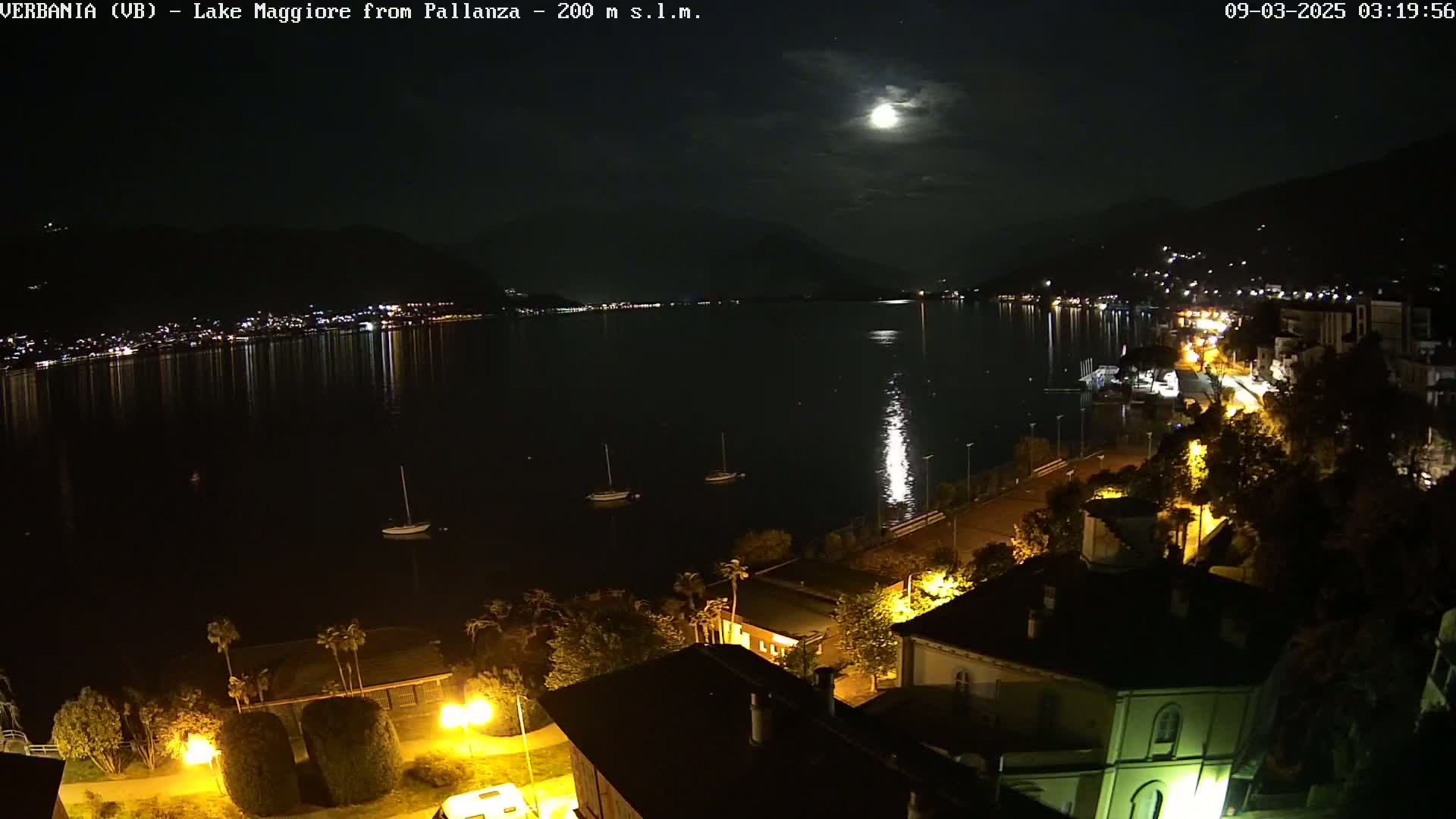 A moonlit night scene shows a calm lake reflecting lights from a town on its shore, with a few boats visible on the water and streetlights illuminating buildings and trees along the lakeside.