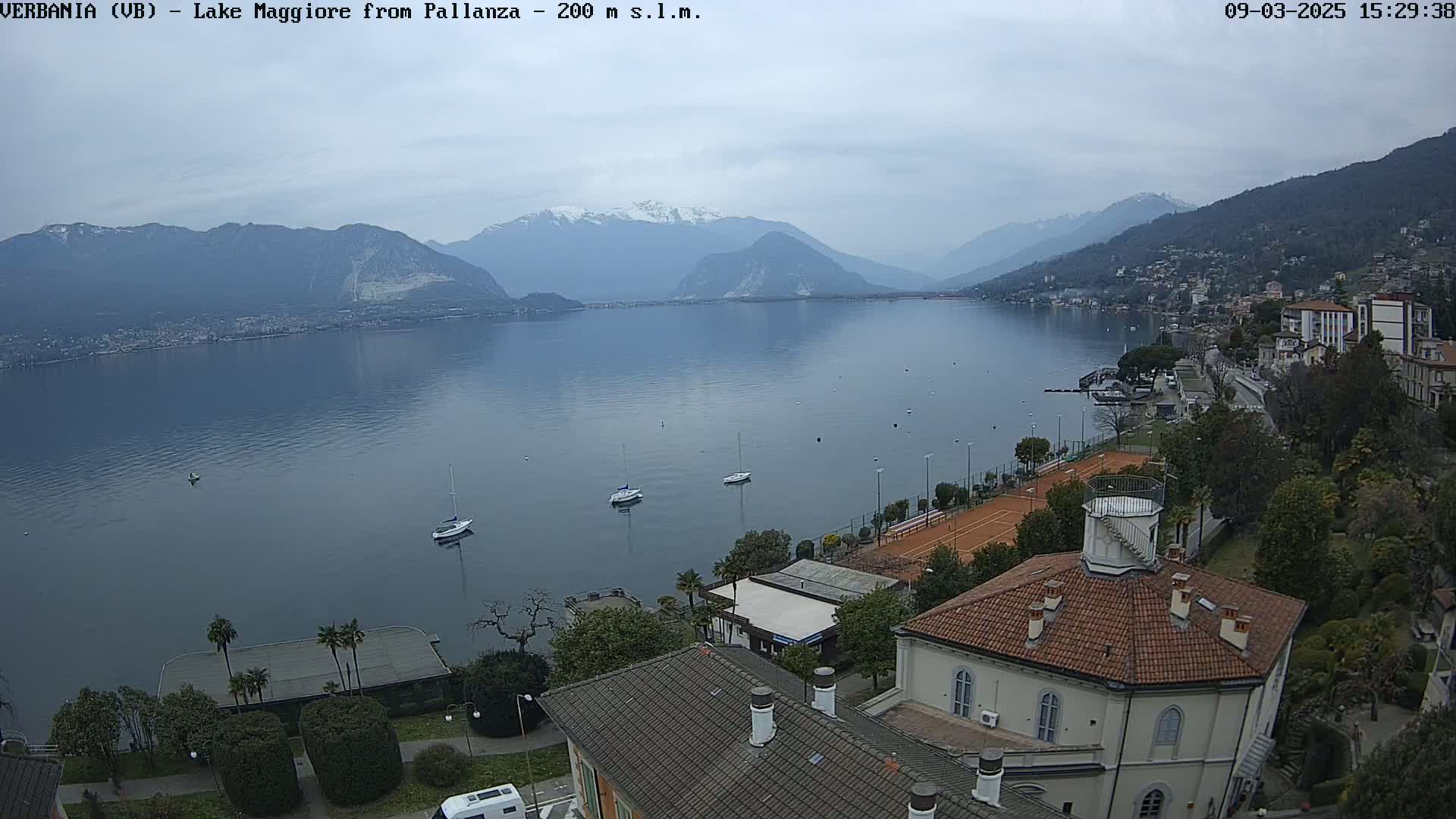 A calm lake stretches to snow-capped mountains under an overcast sky, with several sailboats moored near a lakeside town featuring buildings, tennis courts, and lush greenery.