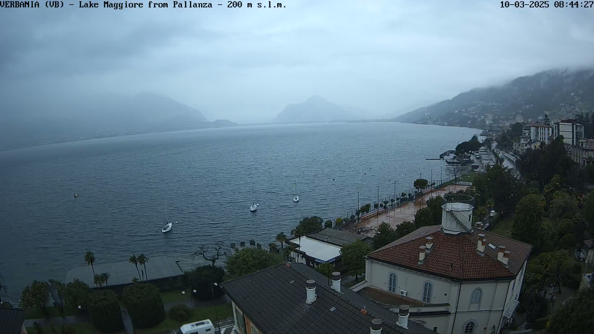 A hazy, overcast day reveals a lake with several sailboats, a shoreline with buildings and trees, and mountains in the distance.