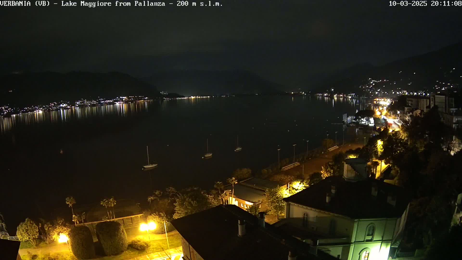 A nighttime view of a calm lake reflecting city lights, with several boats moored near a shoreline lined with buildings and trees under a clear sky.