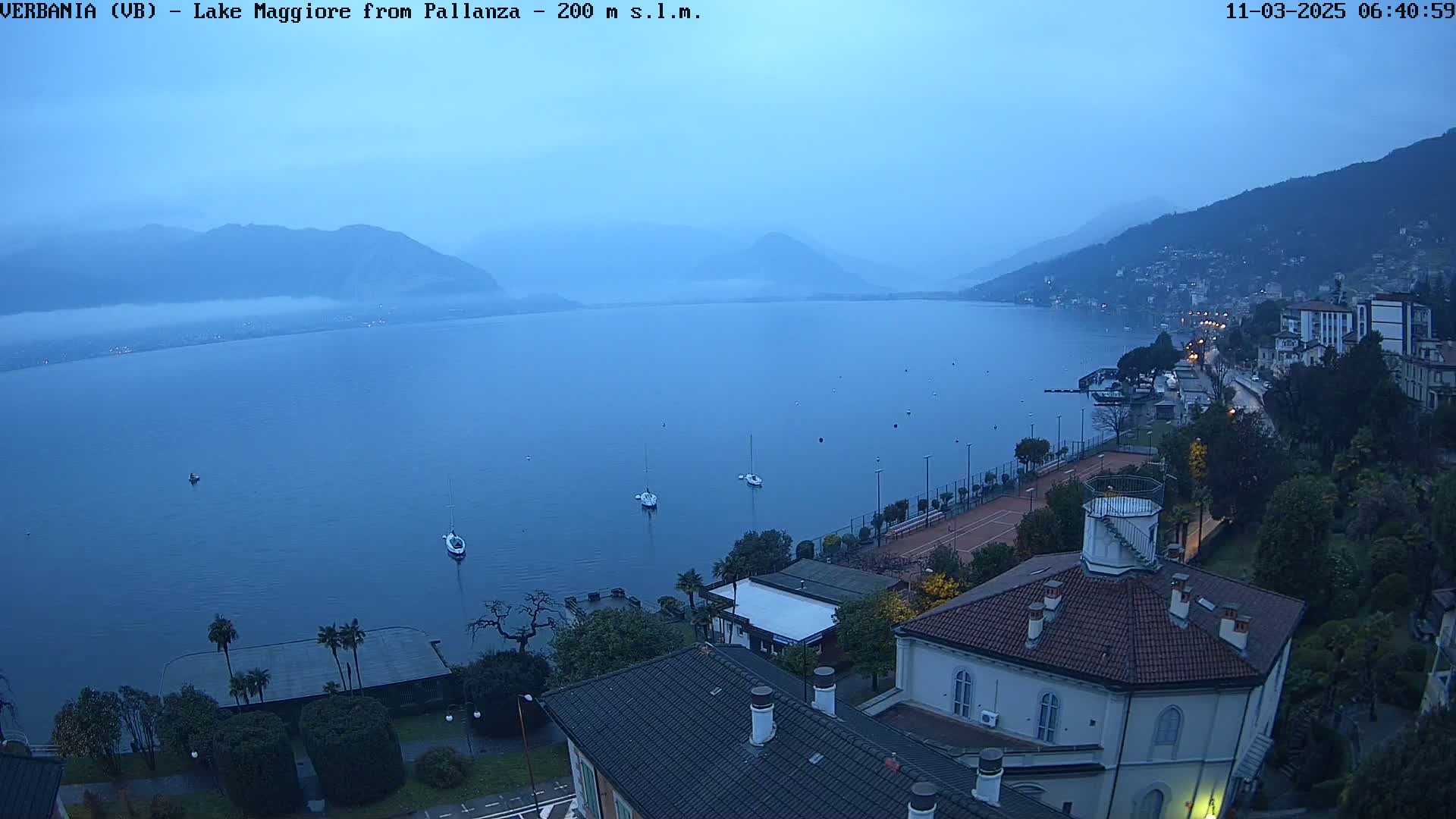 A tranquil lake is seen at dawn, under a hazy sky, with several boats on the water and a town nestled along the shore, featuring buildings and trees.