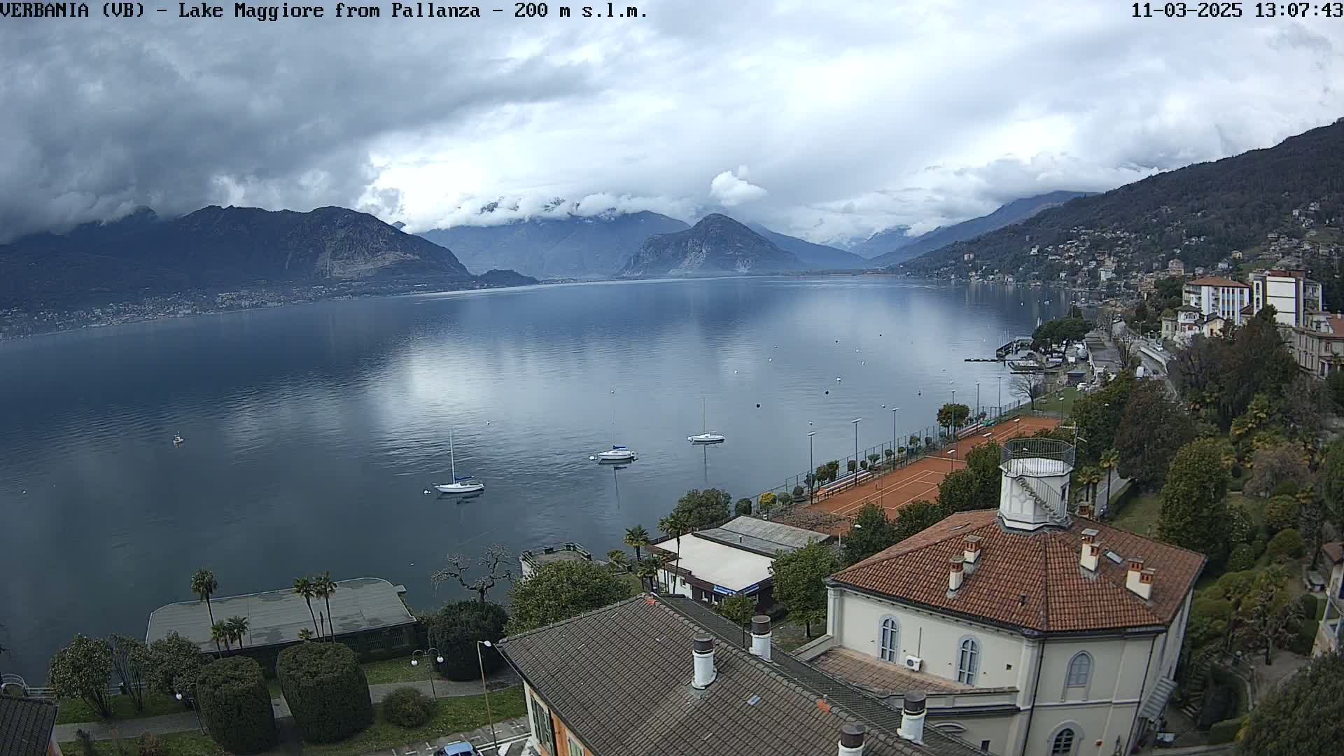 A calm lake reflecting cloudy skies is surrounded by mountains and buildings, with several sailboats at anchor near a shoreline tennis court.