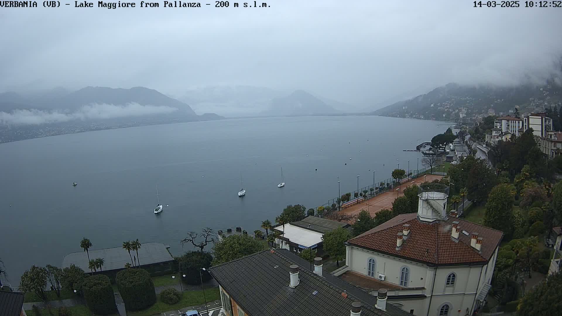 An overcast day shows a calm lake with a few sailboats, a shoreline with buildings and a tennis court, and mountains shrouded in fog in the distance.