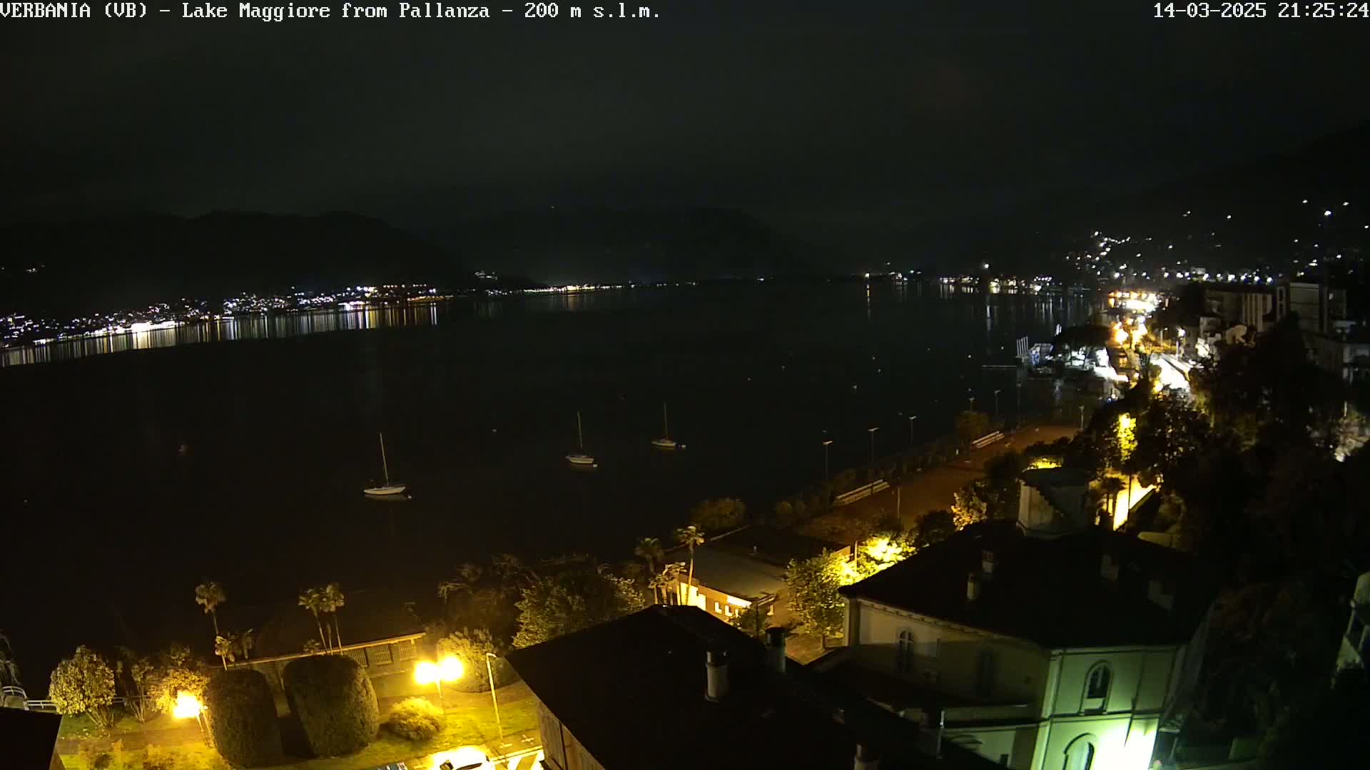 A nighttime view of a calm lake reflecting the lights of a city on its shore, with a few boats visible on the water and illuminated buildings and trees in the foreground.