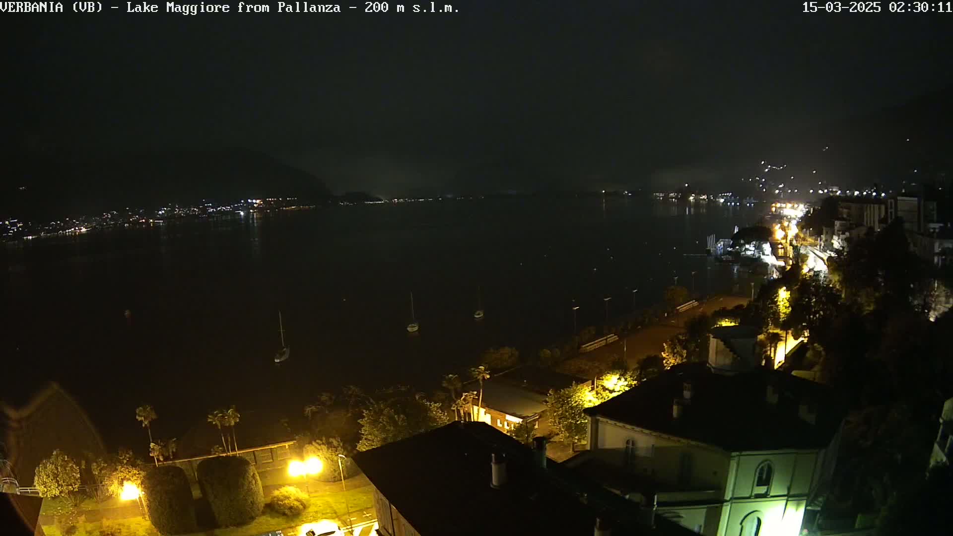 A nighttime view of a dark lake with a few boats, illuminated buildings along a shoreline, and a hazy sky.