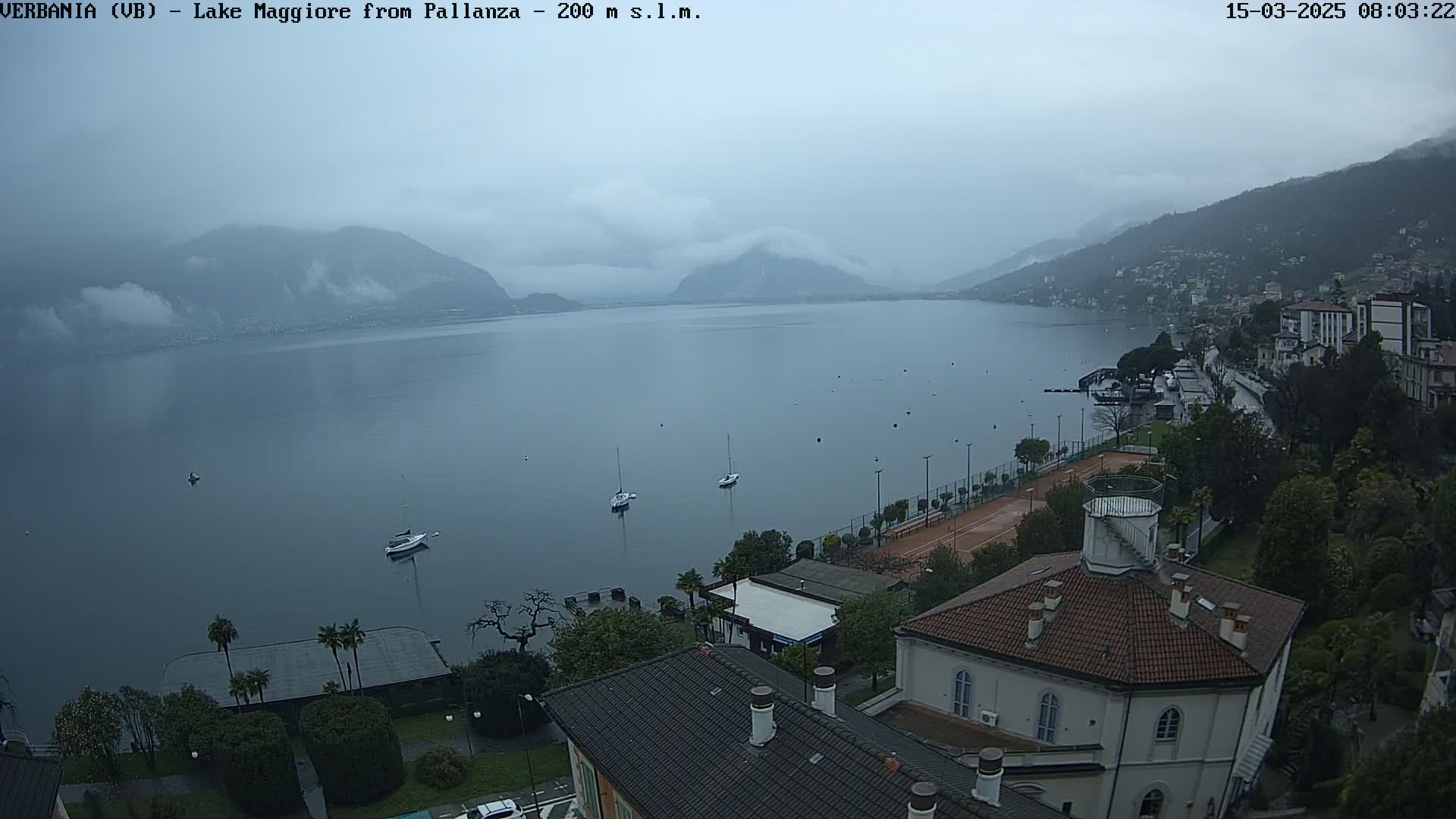 A calm lake with a few boats is seen from an elevated vantage point on a misty, overcast day, overlooking a lakeside town with buildings and trees.
