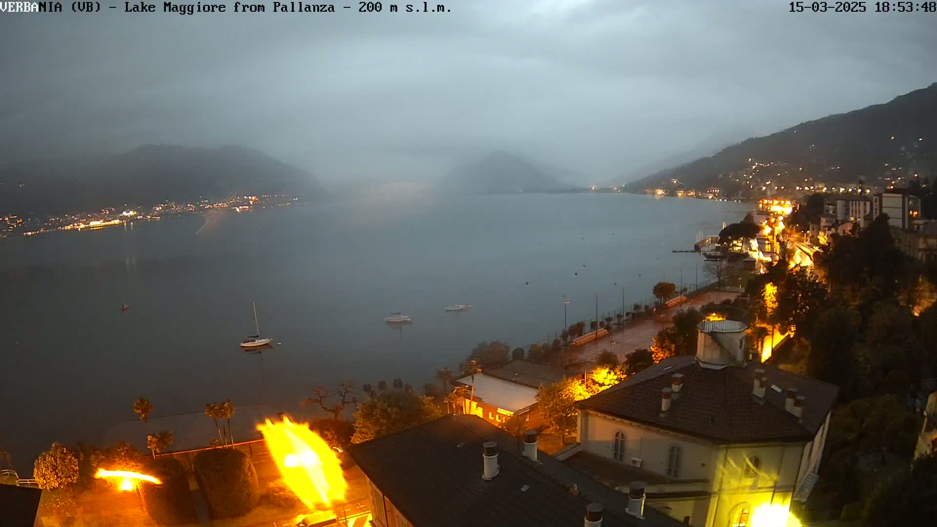 A foggy evening view of Lake Maggiore from a hillside overlooking a town with illuminated buildings and boats on the water.