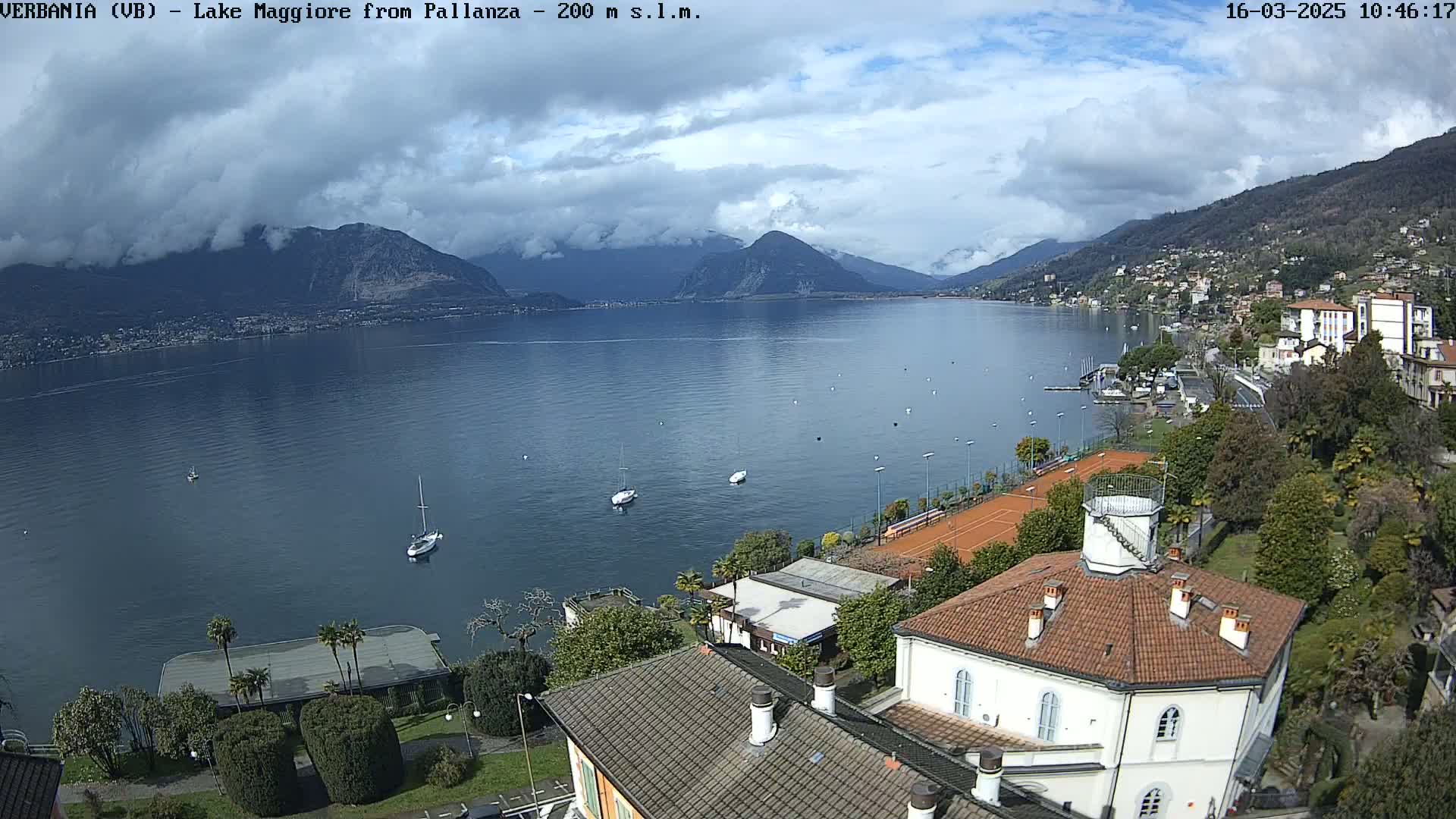 An aerial view shows a partly cloudy day over Lake Maggiore, with sailboats on the water, tennis courts, and buildings near the shoreline, all nestled between mountains.
