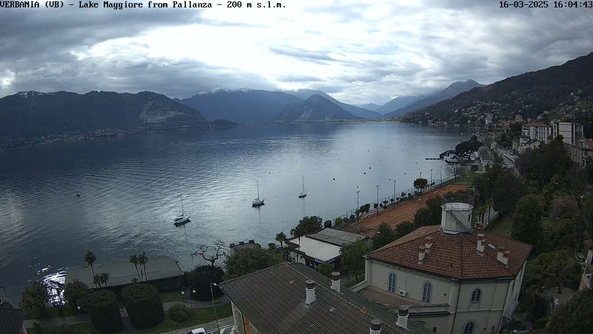 A view of Lake Maggiore from Pallanza, under a cloudy sky, shows sailboats on the water, buildings and tennis courts along the shore, and mountains in the background.