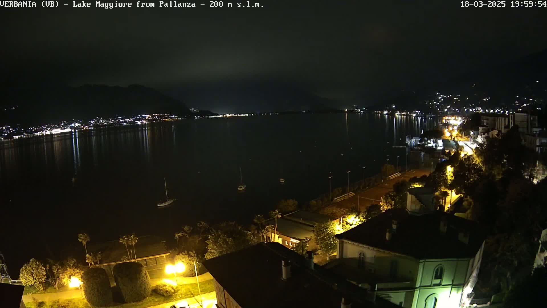 A nighttime view of a calm lake reflecting city lights, with a shoreline featuring buildings and trees illuminated by streetlights.