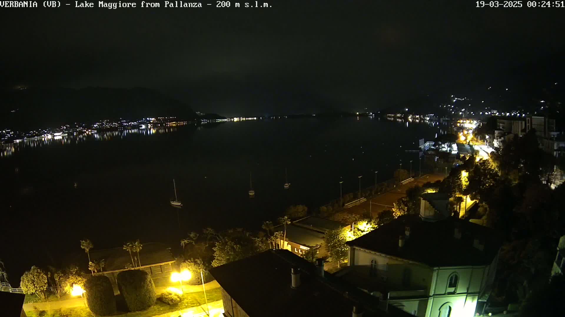 A nighttime view of a calm lake with a town's lights reflecting on the water, seen from a hilltop overlooking buildings and streets illuminated at night.
