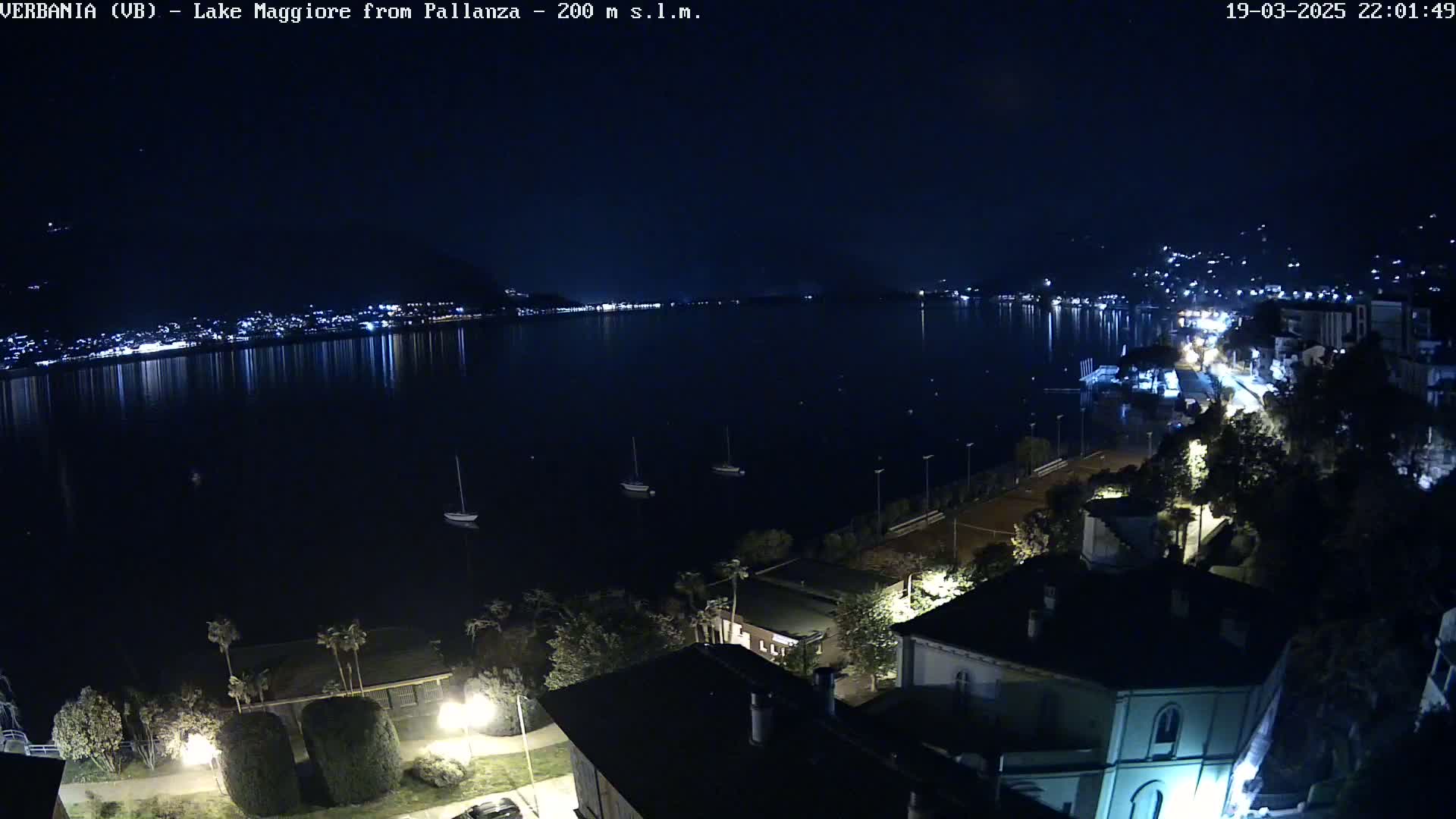 A nighttime view of a calm lake reflecting city lights, with several boats anchored near a shoreline featuring buildings and trees under a clear, dark sky.