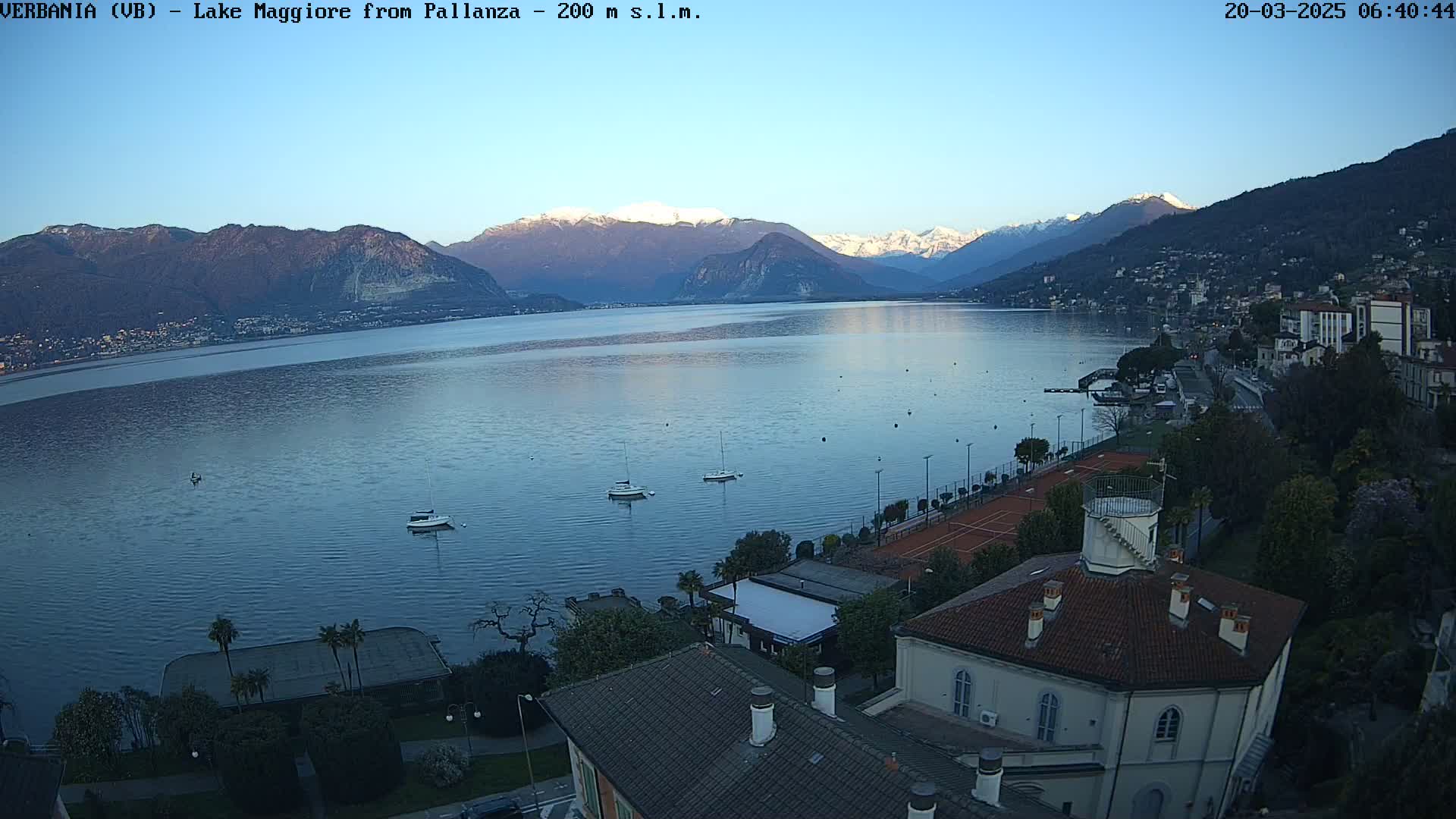 A calm lake with snow-capped mountains in the background, under a clear sky, is viewed from above a lakeside town with several buildings, tennis courts, and a few boats on the water.