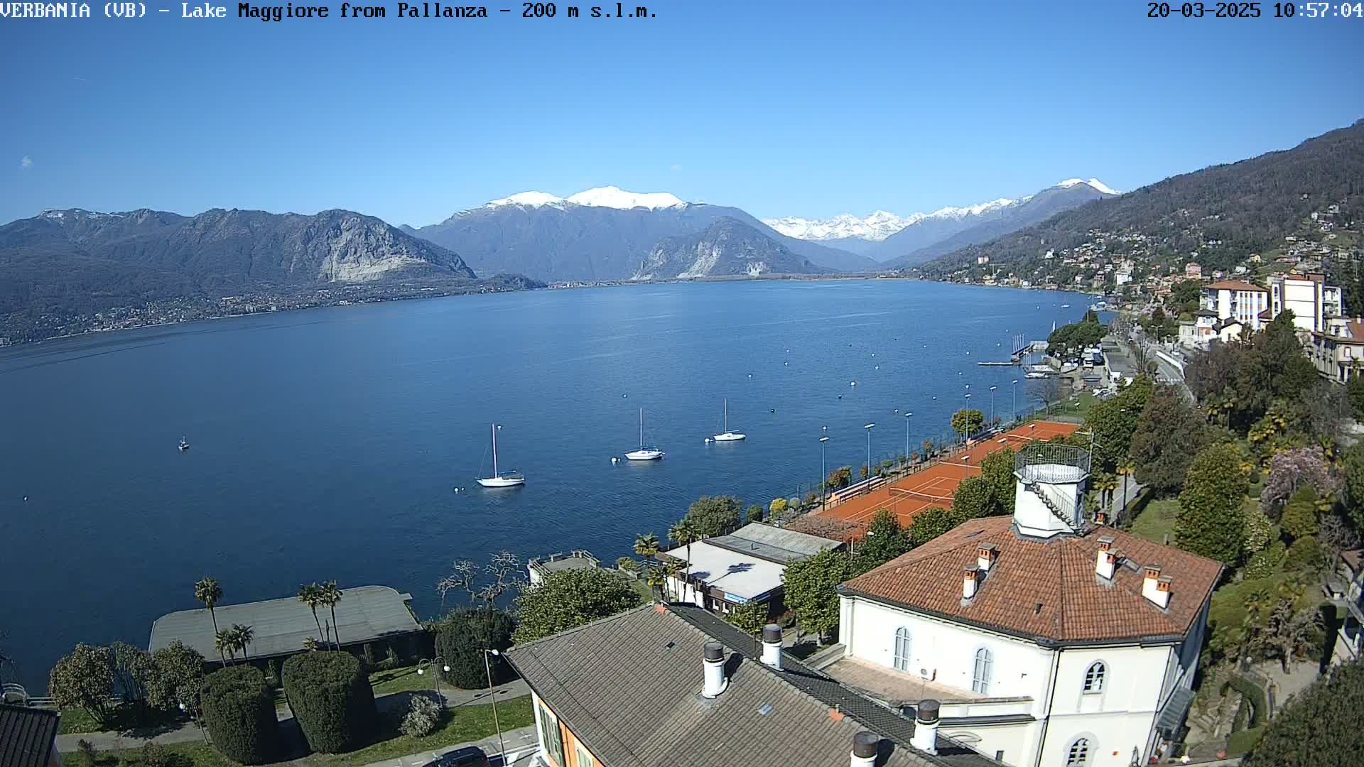 A clear, sunny day overlooks a calm lake with several sailboats, tennis courts, and buildings nestled along the shoreline, backed by mountains with some snow-capped peaks.