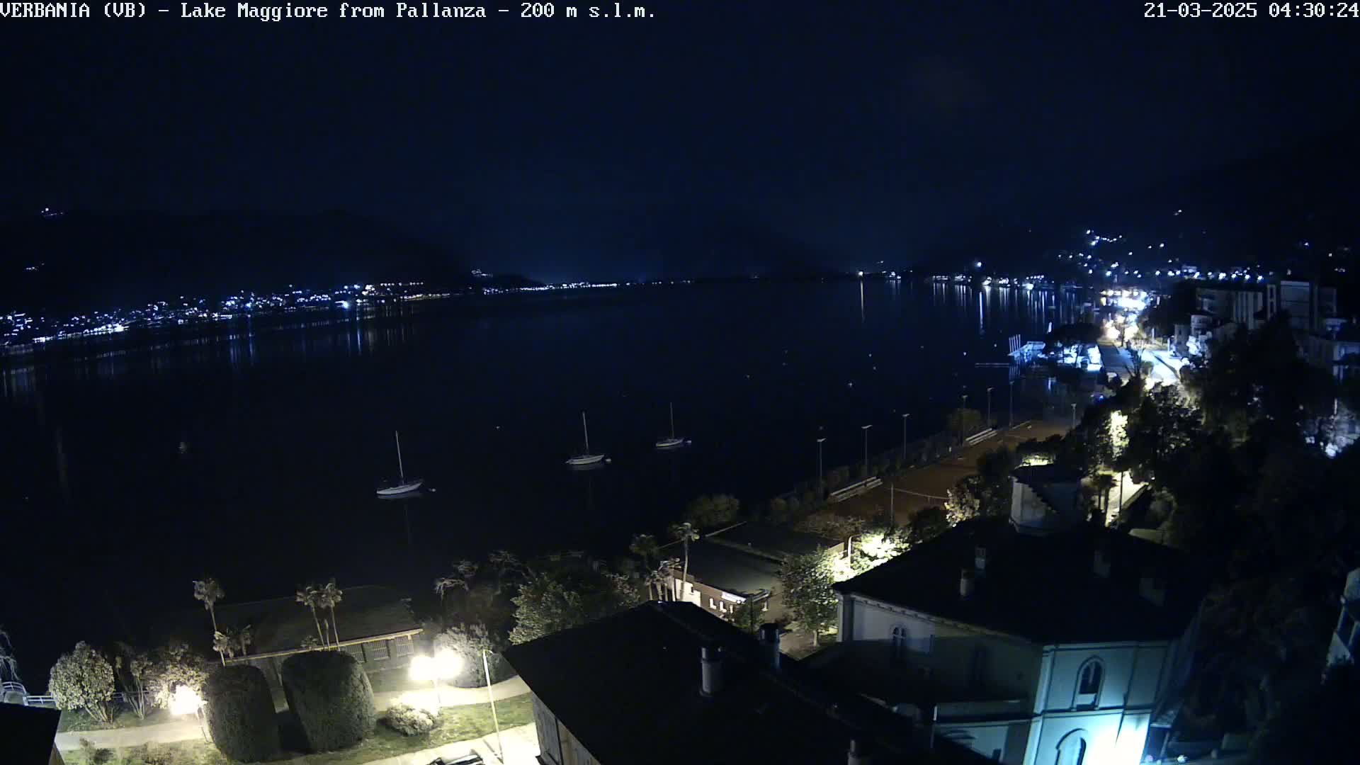 A nighttime view of a calm lake with a few boats, a town with lights on the far shore, and buildings and trees in the foreground under a clear, dark sky.