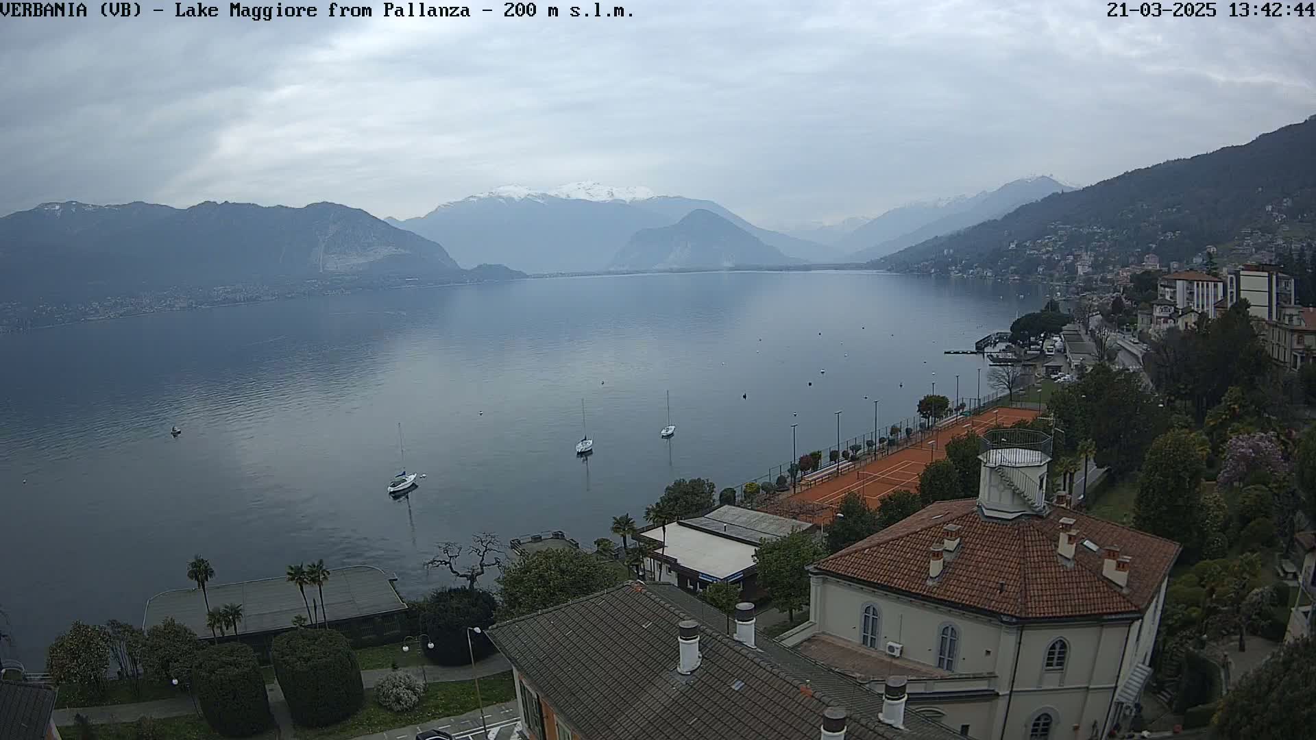 A view of Lake Maggiore from Pallanza on an overcast day, showing several sailboats on the lake, a tennis court, and buildings along the lakeshore.
