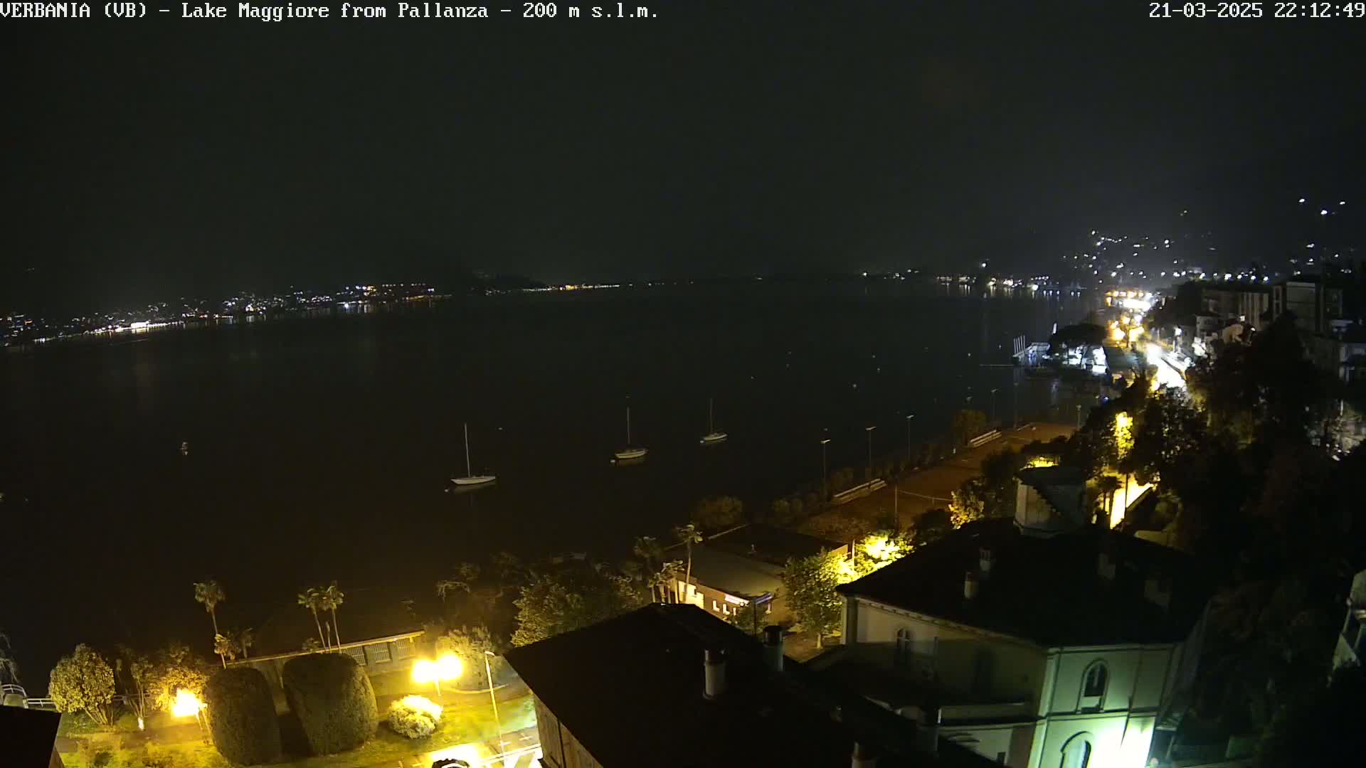 A nighttime view of a calm lake with a few boats, illuminated buildings along the shore, and dimly lit streets.