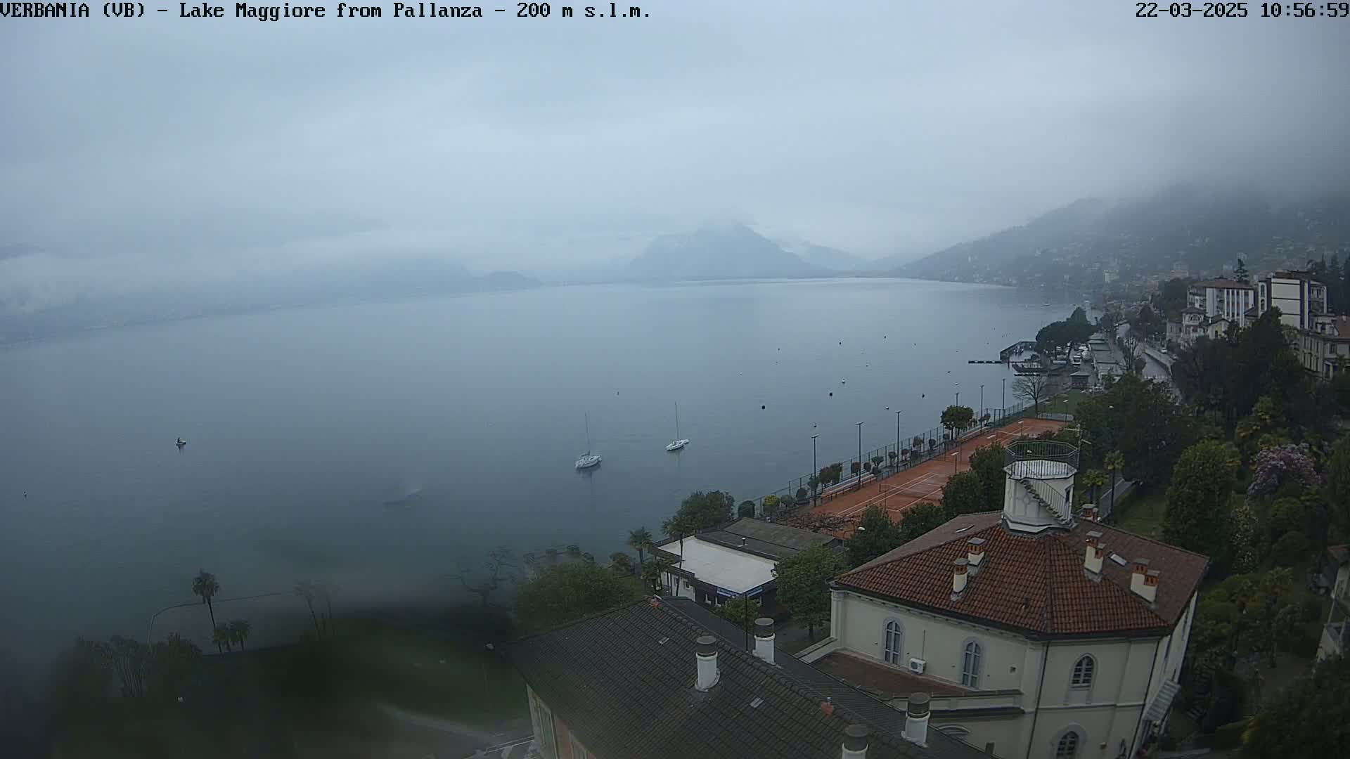 A hazy, overcast view of Lake Maggiore from Pallanza shows a calm lake with a few boats, a tennis court, and buildings along the shoreline.