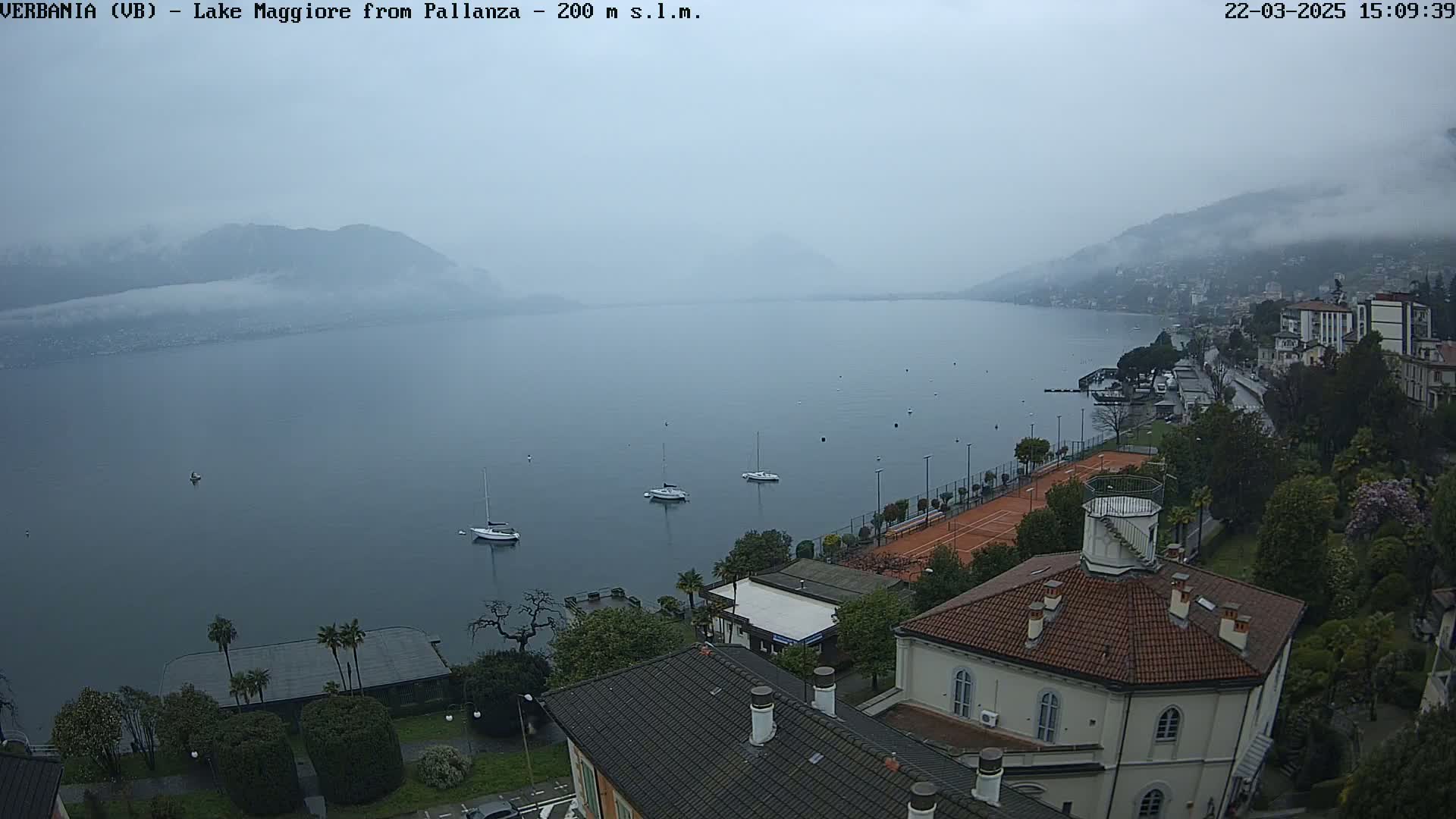 A hazy, overcast day reveals a large lake with several sailboats, a tennis court, and buildings nestled along its shore, all surrounded by greenery.