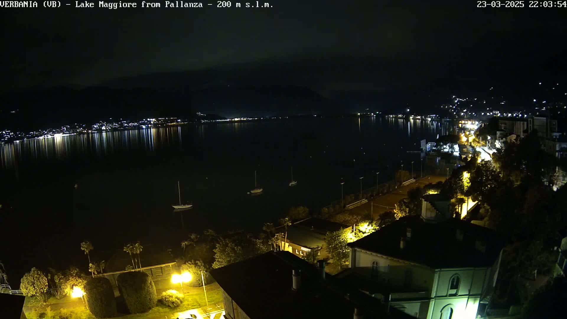 A nighttime view of a lake with a town's lights reflected in the water, boats moored, and buildings illuminated along the shoreline.