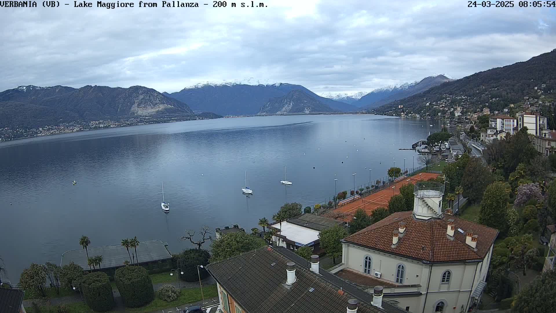 A calm lake stretches out beneath a cloudy sky, bordered by snow-capped mountains and a lakeside town with tennis courts and buildings.