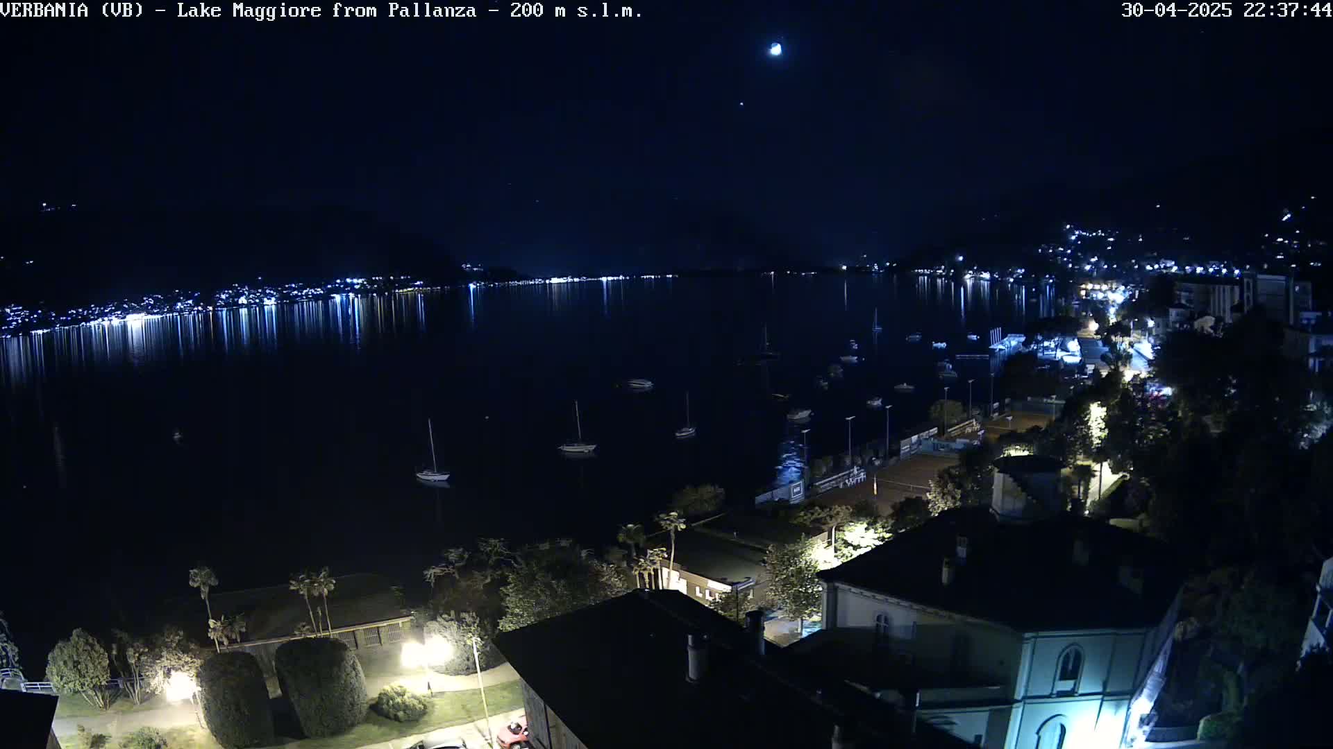 A nighttime view of a calm lake with lights reflecting on the water, boats moored in the bay, and a town with illuminated buildings on the shore under a clear, dark sky.