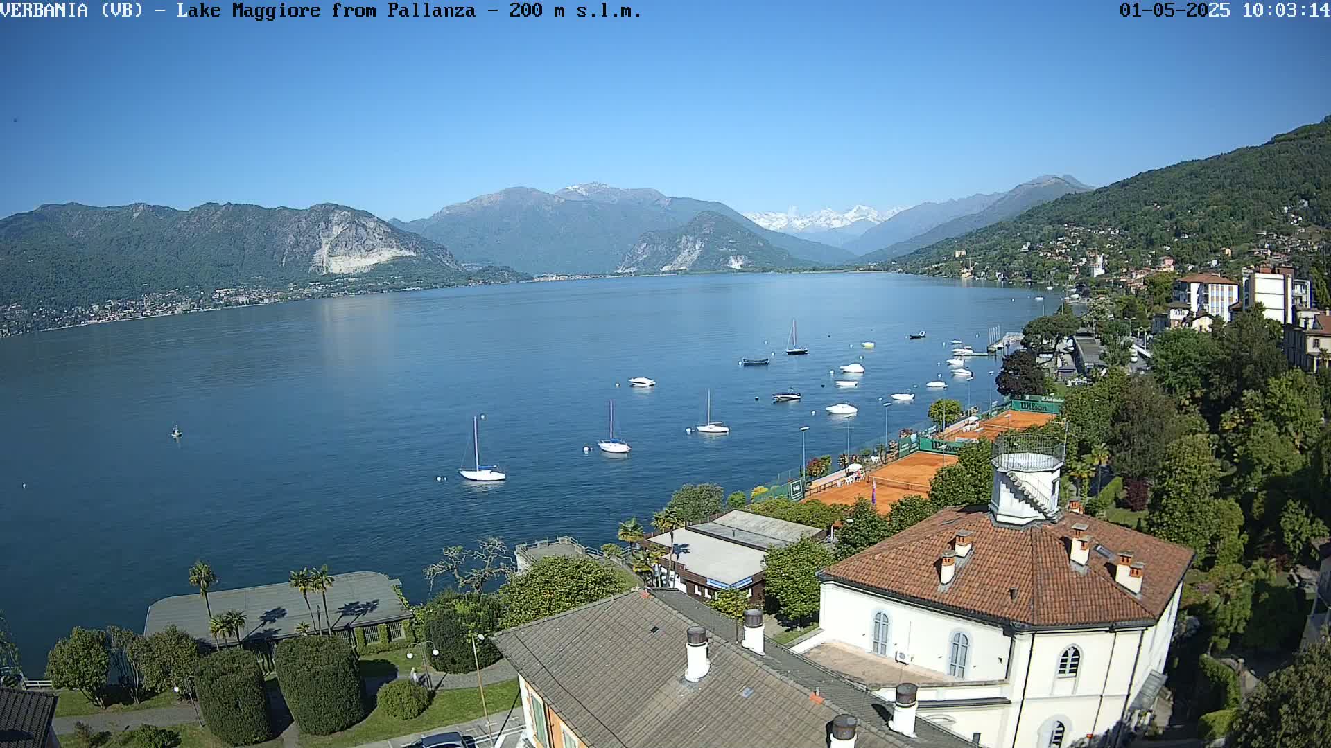 A sunny day overlooking Lake Maggiore, numerous sailboats are moored near a shoreline with buildings and tennis courts, mountains are visible in the background.