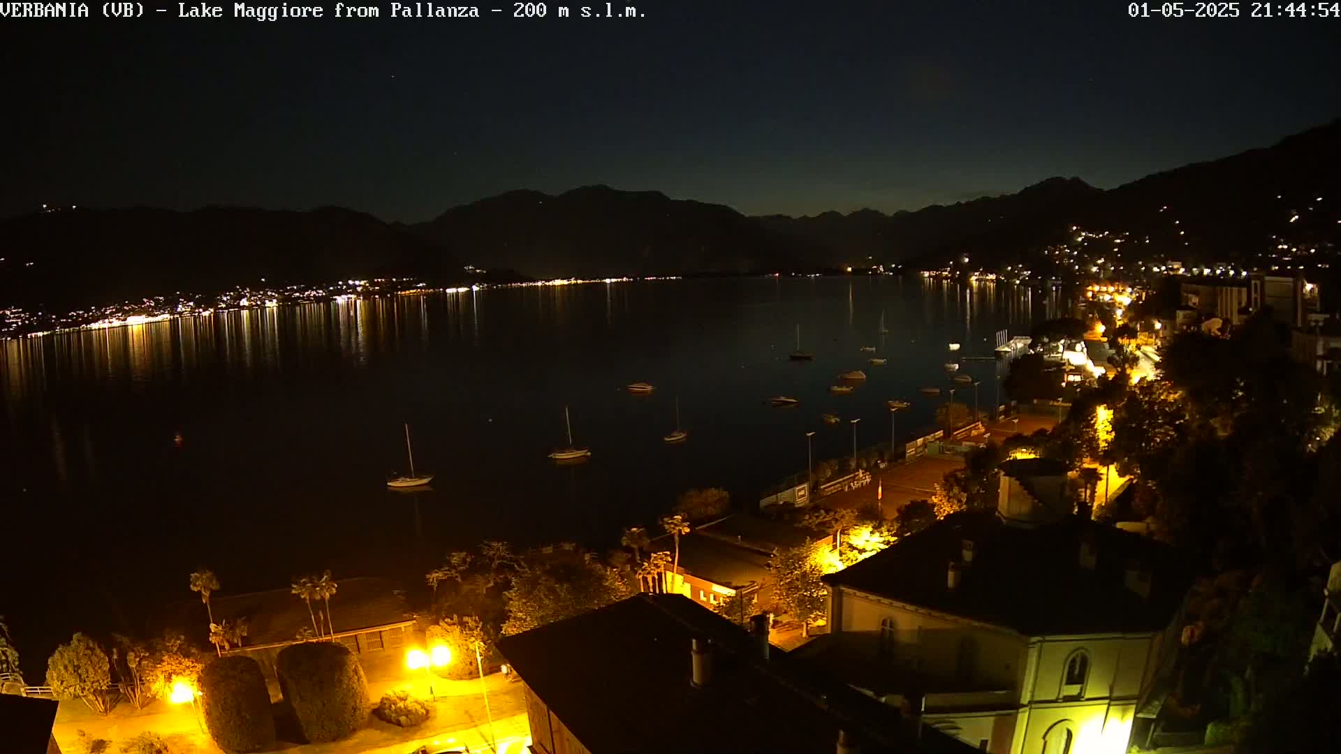 A nighttime view of a calm lake with boats and a town illuminated by streetlights, under a clear, dark sky.